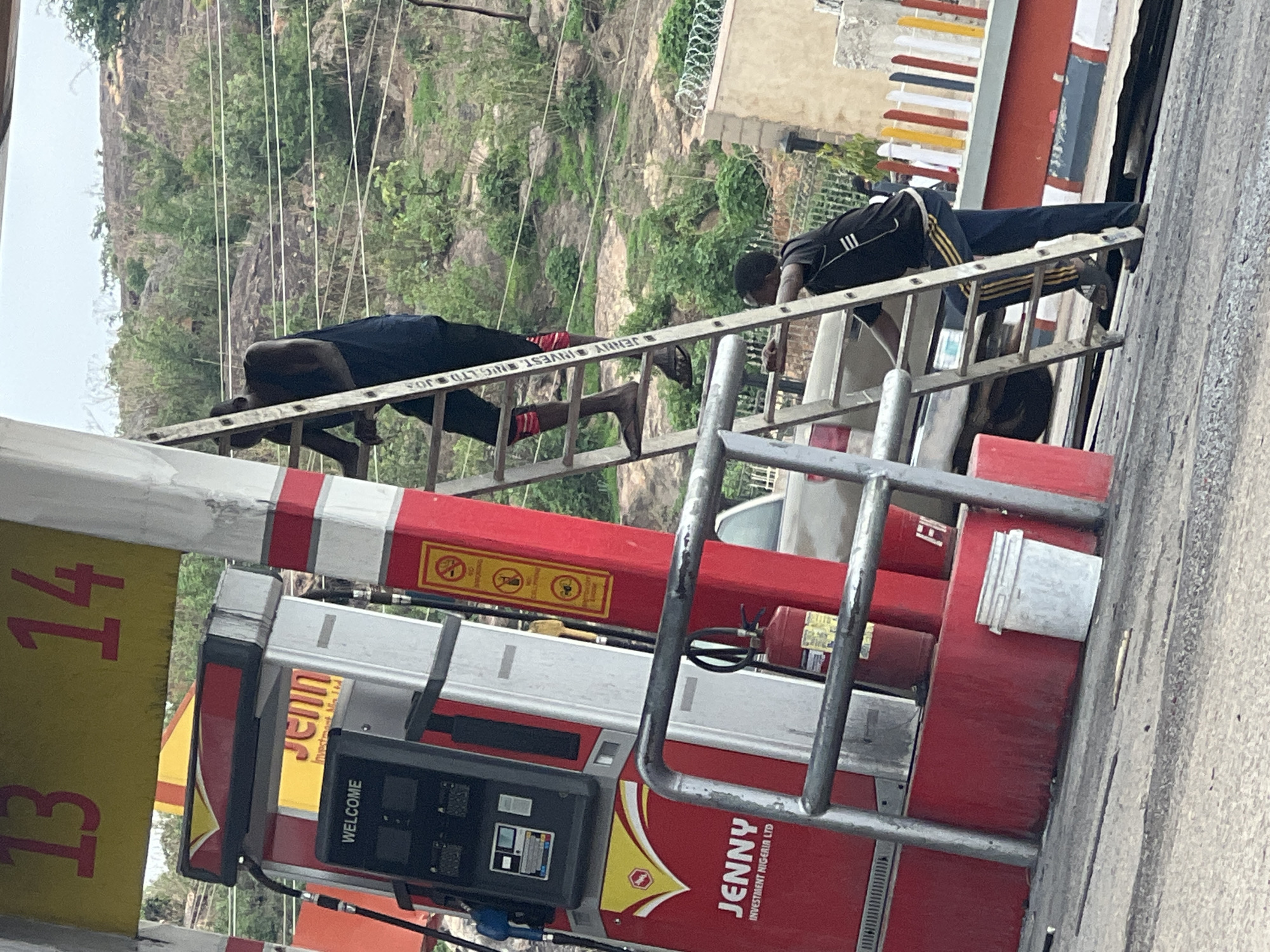 The image depicts a scene in Gura Top, Nigeria, where two men are positioned on a ladder adjacent to a JENNY-branded gas pump. The pump, predominantly red and yellow, displays the number "13.14" on a sign. One man, wearing a dark outfit with red accents on his lower legs, is near the top of the ladder. The other man, in a black athletic-style shirt with white stripes on the sleeves and dark pants, is further down the ladder. The ladder is leaned against the side of a building or structure, and the background features a landscape with vegetation and power lines.