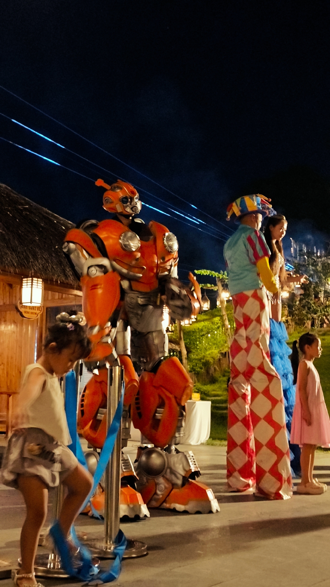 A group of performers, including a person in a large orange robot costume, a stilt walker in a jester-like outfit, and two young girls, are gathered outdoors at night. The robot, which resembles Bumblebee from the Transformers franchise, is positioned in the foreground. The stilt walker is to the right of the robot and is wearing a blue shirt, yellow sleeves, and red and white diamond-patterned pants, along with a colorful jester hat. One young girl, wearing a light-colored top and a grey skirt, is in front of the robot, interacting with blue ribbons wrapped around a pole. Another young girl, wearing a pink dress, stands further to the right. In the background, there are trees, lights, and structures that suggest an outdoor event or restaurant in Hoa Lư Ward, Vietnam. The ground appears to be paved.