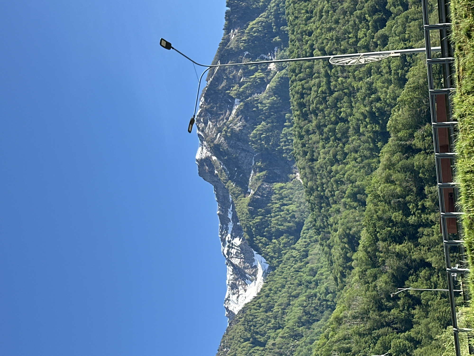 The image displays a majestic mountainous landscape in Krasnaya Polyana, Russia, under a vivid, clear blue sky. A prominent mountain dominates the scene, its slopes covered with dense, vibrant green forests. A significant portion of the mountain face features a steep, rugged rock formation, accented by striking white patches of snow or ice that cascade down its dark surface.
In the foreground, on the right side, a modern street light with a dark pole and a rectangular LED lamp extends into the frame. Further to the right, a multi-tiered metal structure, possibly part of a building facade or a retaining wall, is visible, partially covered by abundant green vegetation, giving it an overgrown appearance. Another, smaller street light fixture is discernible lower down, partially hidden by the trees and the structure. A distinct circular decorative element with spokes is also attached to a pole near the top right, possibly another light fixture or signage.
The scene is illuminated by bright daylight, indicating clear and sunny weather. No people are visible, emphasizing the grandeur of the natural environment. The presence of snow or ice on the mountain face, even under a bright sky, suggests either high altitude or a cooler season. No visible text is present in the image.