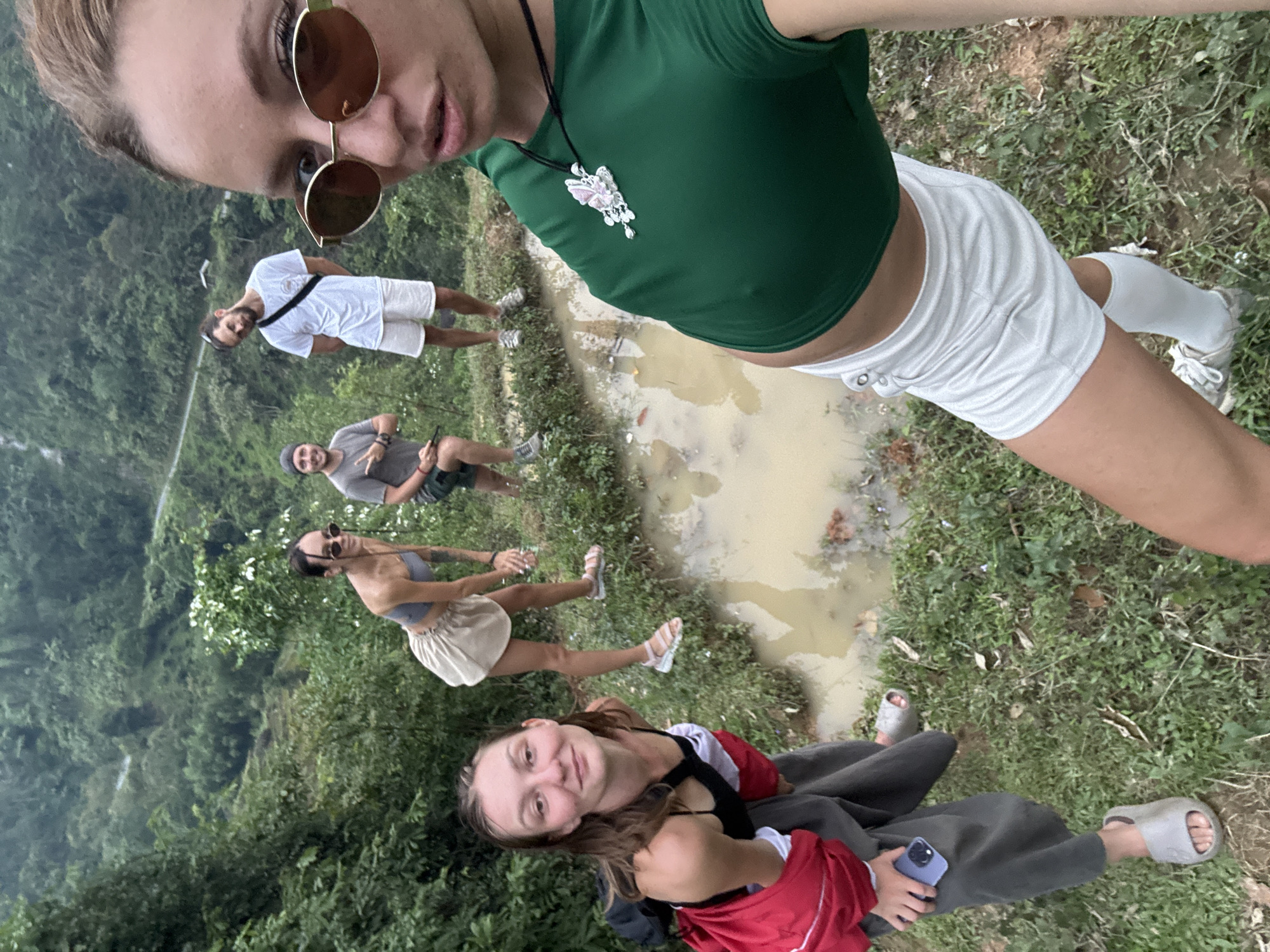 The image is a tilted outdoor selfie taken in Nậm Dịch Commune, Vietnam, featuring a group of five individuals in a natural, lush green environment.
In the immediate foreground, the person taking the selfie is a young woman with light hair, wearing large round brown sunglasses, a dark green sleeveless crop top, and white athletic shorts. She has a silver butterfly pendant necklace.
Behind her, arranged from left to right in the midground:
1. A young man with dark hair and a beard, standing and looking towards the camera, wearing a white t-shirt and white shorts with sneakers.
2. Another young man with dark hair and a beard, wearing a grey t-shirt and shorts, a dark hat, and holding a mobile phone. He is seated on the ground near a muddy puddle.
3. A young woman with dark hair, wearing sunglasses, a grey bandeau top, and a light-colored skirt, standing on the grass.
4. In the closer foreground, another young woman with long dark hair is seated on the grassy ground, looking towards the camera with a slight smile. She is wearing a black top, a red top draped over her shoulders, dark pants, and grey slides, holding a mobile phone in a purple case.
The setting is a verdant landscape with abundant green foliage, suggesting hills or mountains in the background. A large, irregularly shaped puddle of muddy water is prominent in the midground, indicating damp ground conditions. The ground around the puddle is a mix of grass and dirt. The overall scene suggests a casual outdoor gathering, possibly during a hike or travel excursion, under daylight conditions.