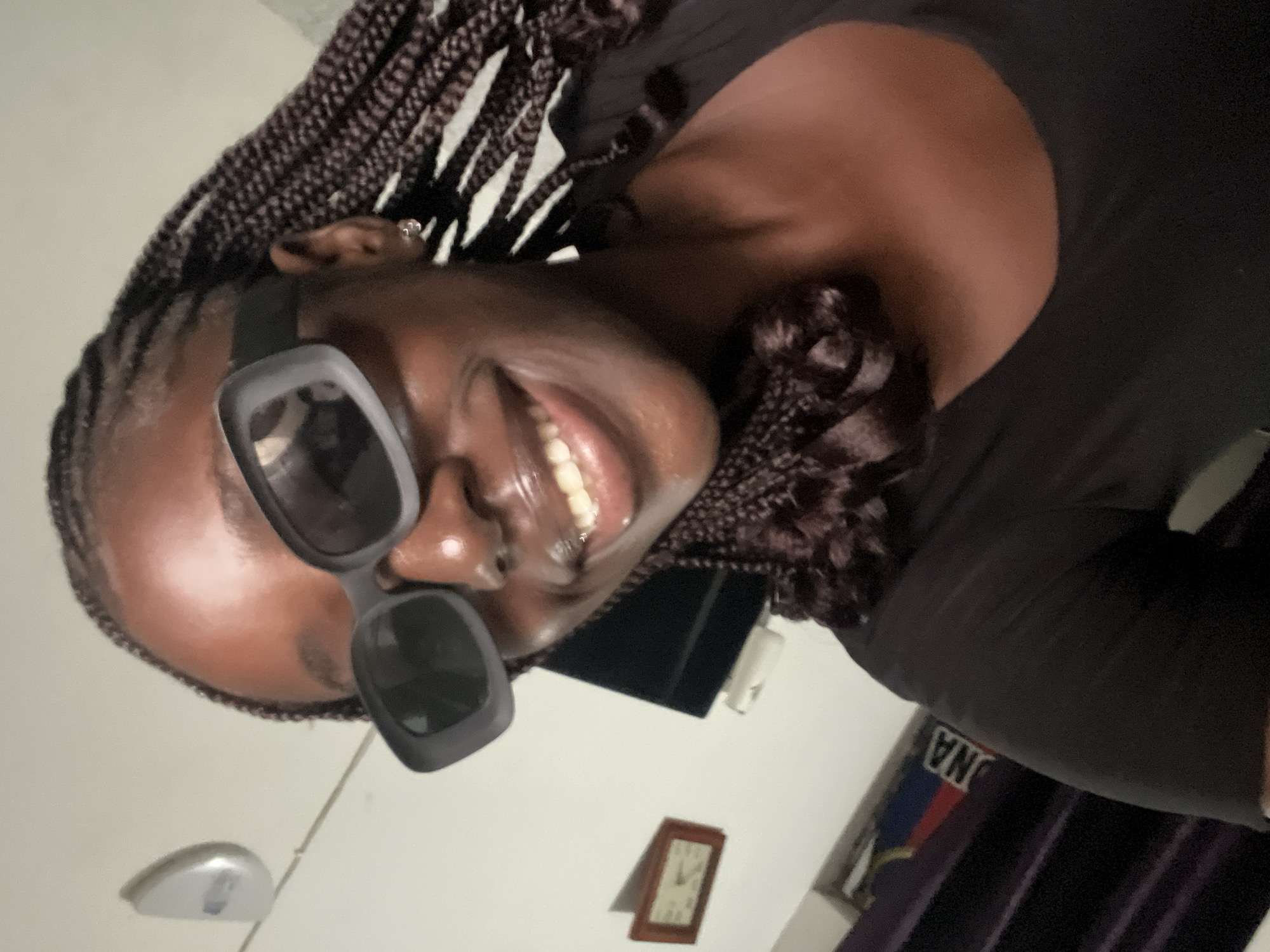 A medium shot looking up at a woman with dark skin, braided hair, and large sunglasses. She is smiling broadly, revealing her teeth. She wears a dark shirt. Behind her, there is a wall, with a clock and other items visible. There is no way to tell the location of the image, as Abuja and Nigeria are not visible.