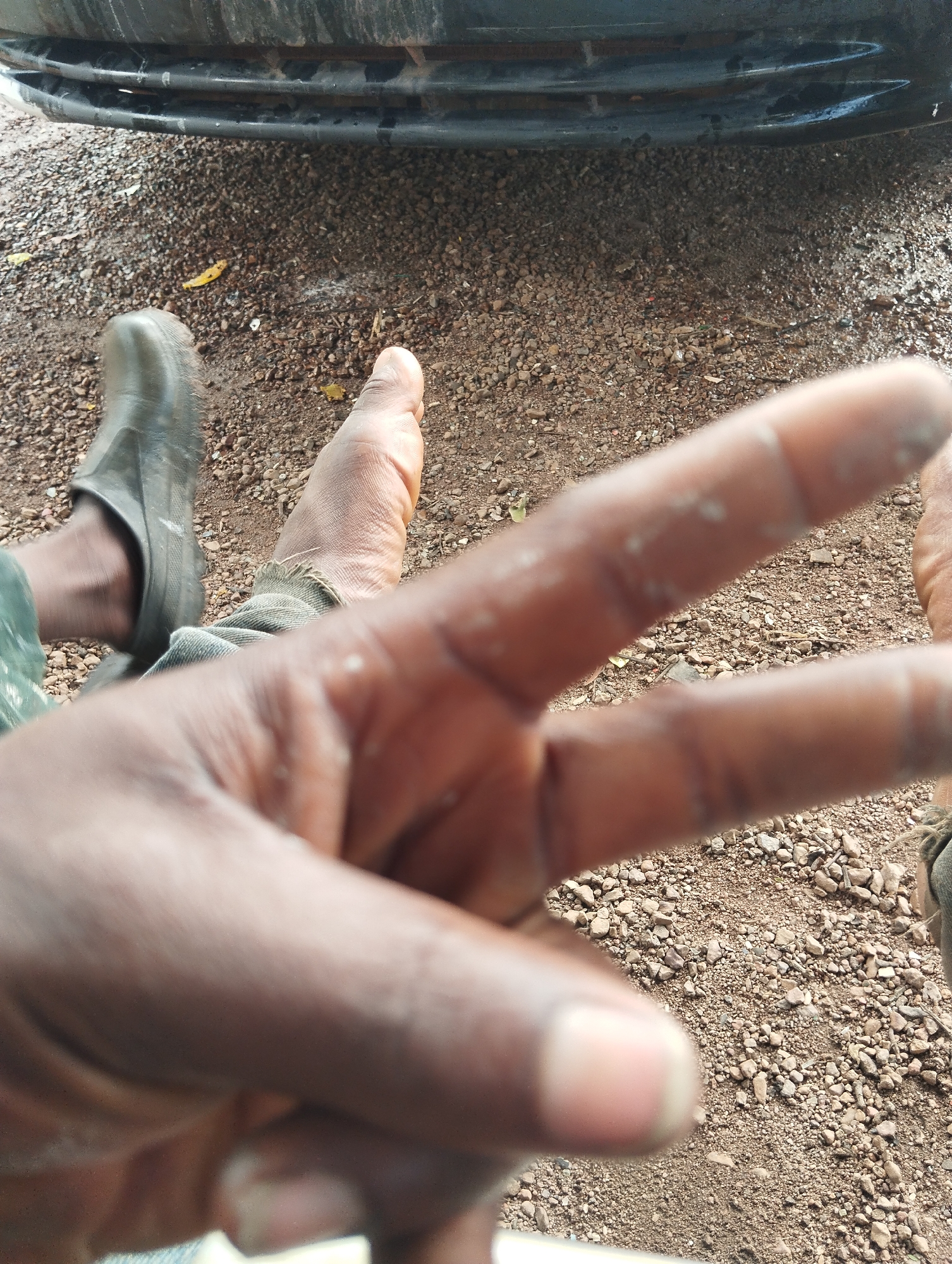 The image shows a person's hand with two fingers extended, forming a peace sign. The hand is in the foreground and appears to be dirty. In the background, there are two feet, one wearing a dark boot and the other bare. The feet are resting on a gravelly ground. Behind the feet, a portion of a vehicle bumper is visible. The scene appears to be outdoors, possibly in an unpaved area. The overall impression is that of a candid snapshot taken in a casual setting. The location of Bukuru, Nigeria is not explicitly depicted or inferable from the visual elements of the image.