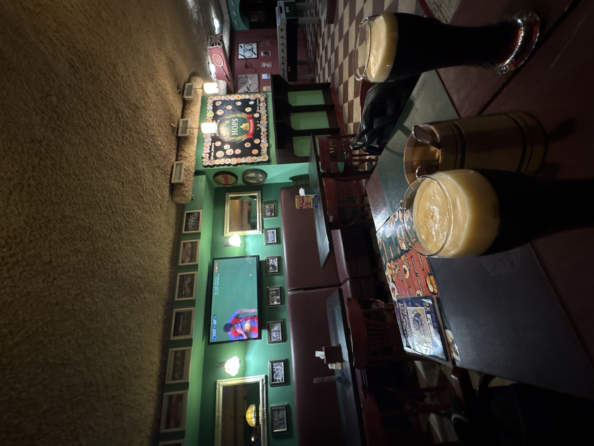 The image depicts the interior of a pub or restaurant in Minsk, Belarus. The walls are green with numerous framed pictures, and a television is mounted on one wall displaying a sports game. There are tables and chairs in the foreground, set with menus and drinks including beer in glasses and a wooden mug. The pub has a distinctive decorative motif, including a sign that reads "HOPS" and unique lighting fixtures. The environment suggests a relaxed, convivial atmosphere.