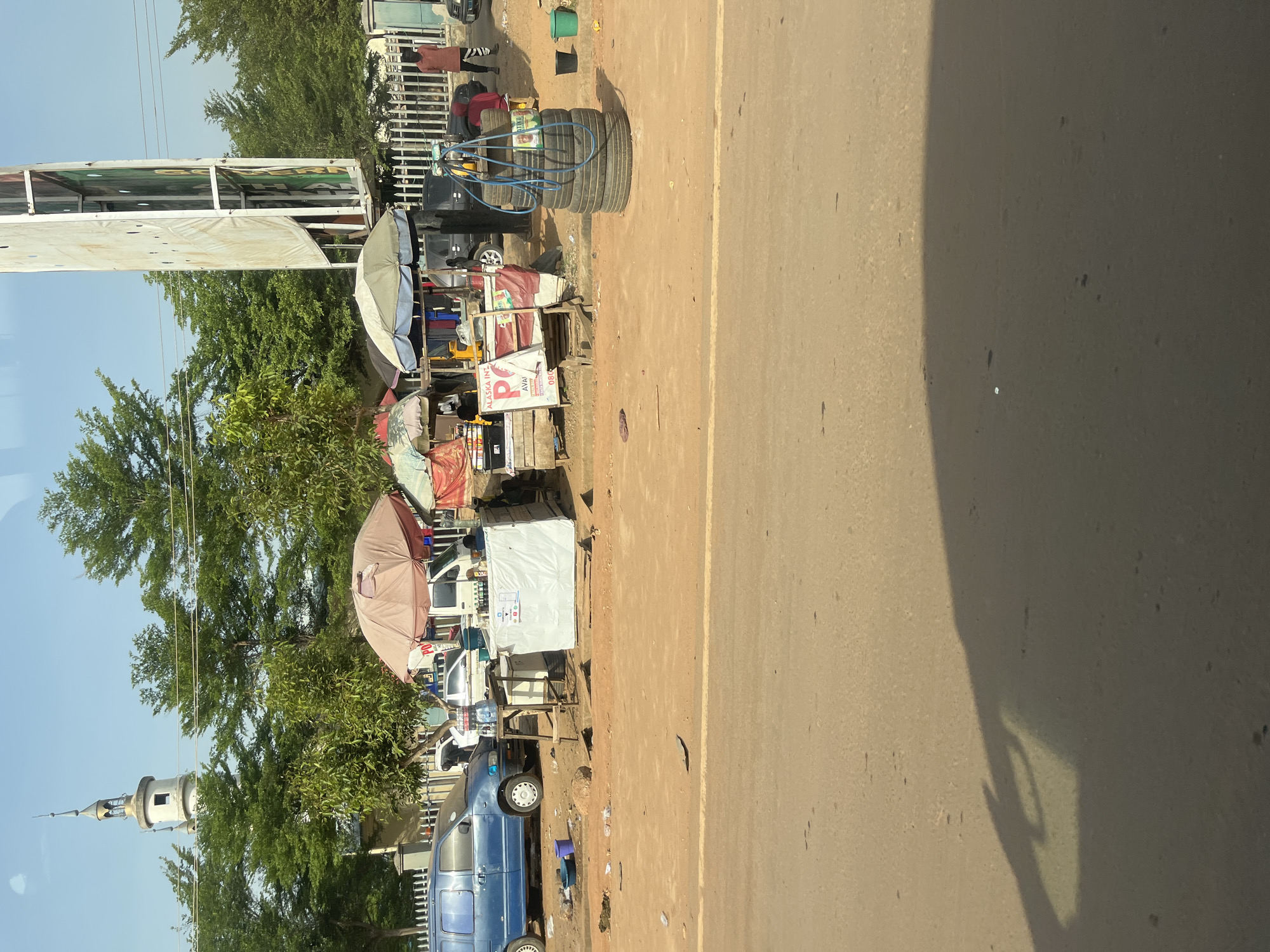 This media file captures a vibrant roadside market scene in Ado, Nigeria, under bright, sunny conditions. The image is composed with a paved road occupying the right half and a bustling market area on the left, set against a backdrop of lush green trees and a clear blue sky.
The main subjects are the various makeshift stalls and vendors operating along what appears to be an unpaved shoulder or dirt area adjacent to the road. Several large umbrellas, in shades of light gray/white and brown/maroon, are set up to provide shade over the stalls and merchandise. Goods displayed include a prominent stack of black vehicle tires with blue tubing wrapped around them, suggesting a tire repair or sales service. Nearby, colorful suitcases and bags (blue, yellow, red) are arranged for sale. Other stalls feature white tarpaulin covers, and some have plastic bottles of water available. A blue van or minibus is parked among the stalls, and other vehicles are partially visible in the background.
A few indistinct figures are present near the stalls, suggesting active commerce, though their specific activities are not clearly discernible. In the far background, a distinctive white minaret-like structure with a pointed spire rises above the tree line, possibly indicating the presence of a mosque nearby.
Notable details include the strong, well-defined shadows cast across the road and from the umbrellas, indicating a bright and sunny day, likely mid-morning or afternoon. The road itself appears to be paved and has a lighter-colored line down its center. A clear shadow of a hand, possibly holding a phone, is visible on the road in one of the crop images, suggesting the photo was taken from a moving vehicle or while standing close to the road.
Visible text on a white sign with a red logo includes "ALASKA INT...", a prominent "P", "AVAI..." (likely "AVAILABLE"), and a partial phone number "080...". This signage points to specific businesses or products being offered. The overall scene depicts everyday life and commerce in an informal outdoor setting typical of the region.