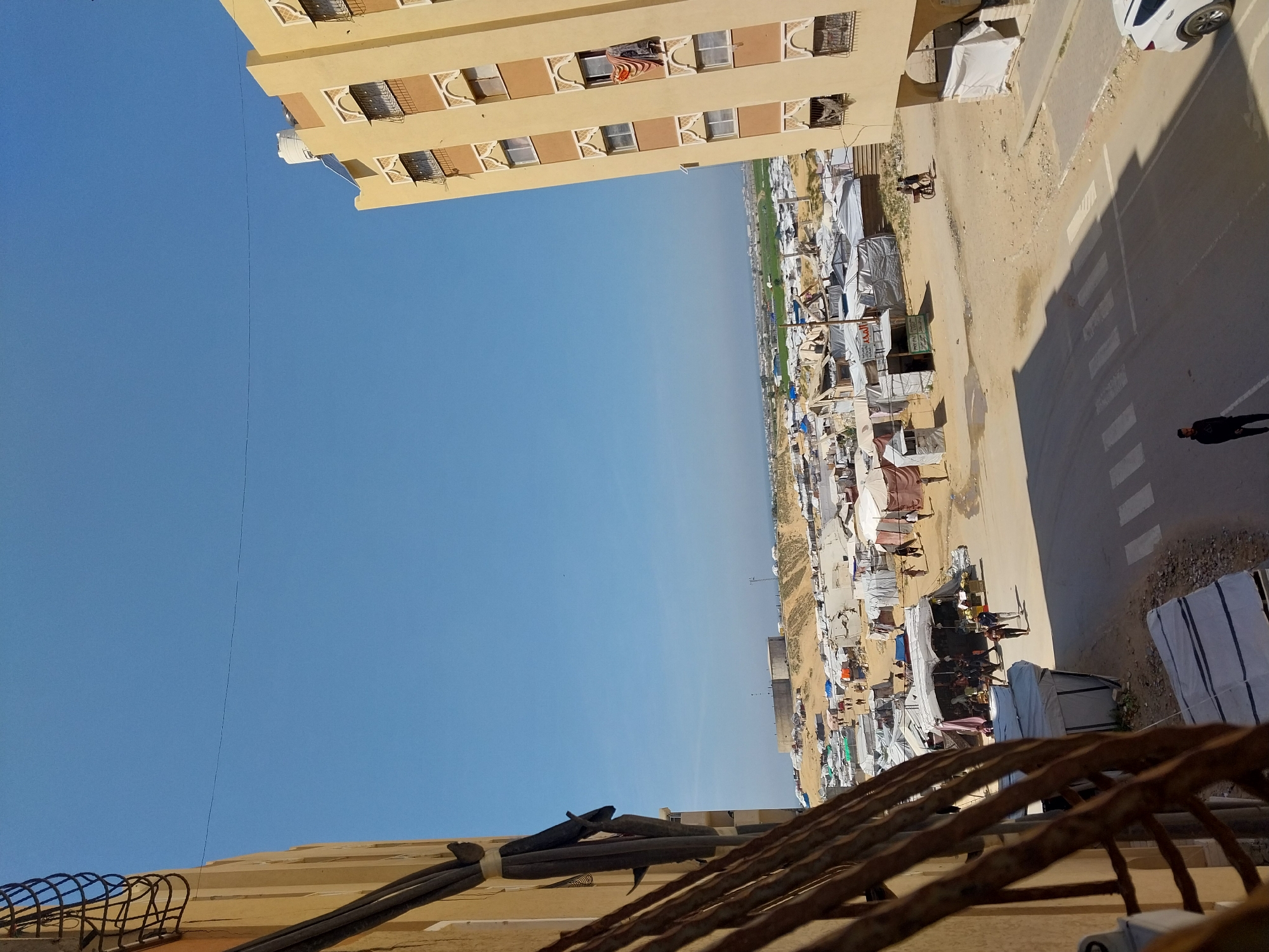 The image shows an aerial view of a building and what appears to be a refugee camp. The top left corner features a multi-story building with windows and balconies. Below this is the camp, consisting of numerous tents or shelters closely packed together. A road runs alongside the camp, with a pedestrian crossing and a few vehicles. A man is walking on the sidewalk. In the foreground is a portion of a building with cables and a metal fence. The sky is clear and blue. The location is Unknown, Unknown.