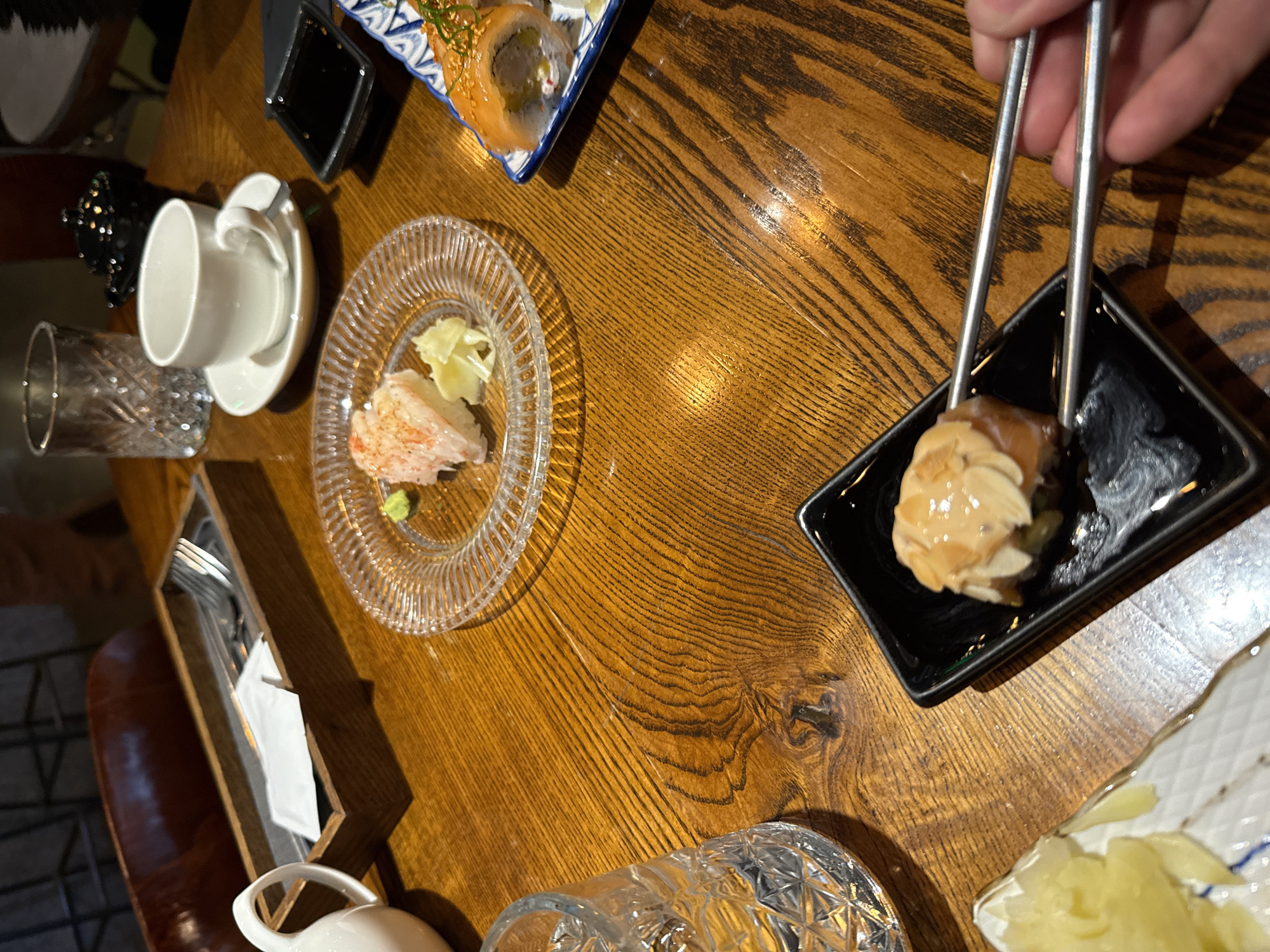 The image depicts a close-up view of a wooden table at a sushi restaurant in Saint Petersburg, Russia. On the table are various sushi dishes, chopsticks, a white cup and saucer, a glass of water, and a small black soy sauce dish. In the foreground, a hand is using chopsticks to pick up a piece of sushi. The sushi appears to be topped with sliced almonds and a creamy sauce. In the background, a white teapot and a dark, lidded container are visible. The overall lighting suggests an indoor setting, possibly in the evening.