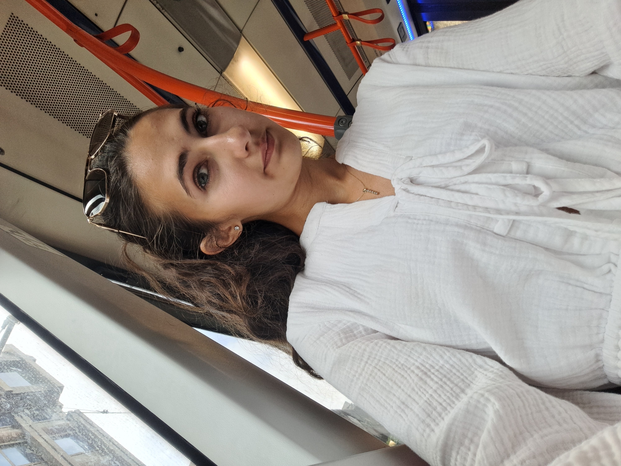 A woman with long, dark, wavy hair is taking a selfie while sitting inside a train or bus. She is wearing a white, loose-fitting, button-up shirt. She has on sunglasses perched on top of her head and a delicate necklace. Her expression is neutral, and she is looking directly at the camera. The interior of the vehicle features orange handrails and beige textured panels. Outside the window, a blurry view of buildings can be seen, suggesting an urban environment. The lighting suggests it is daytime.