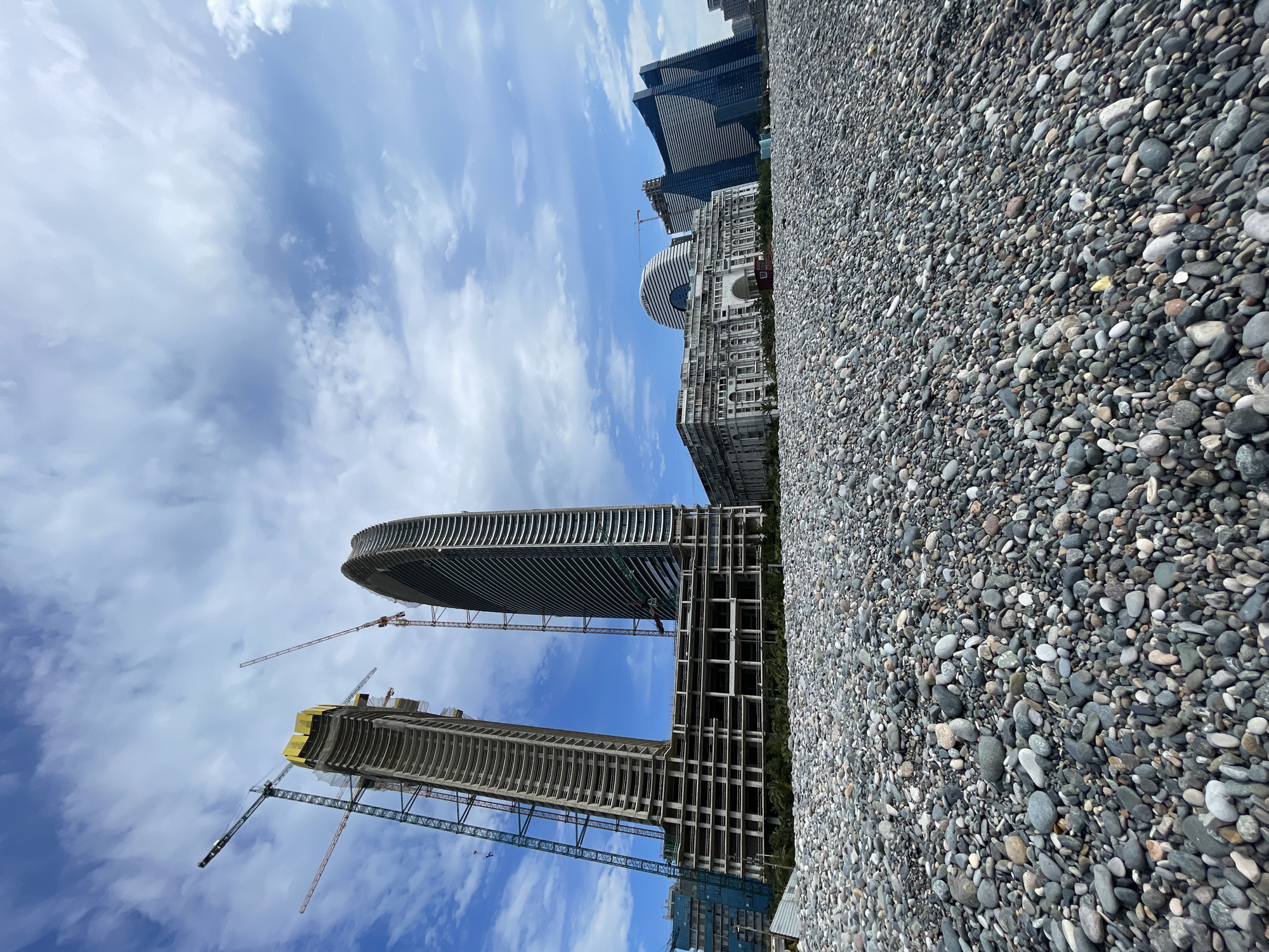 A low-angle shot depicts a pebble beach in Batumi, Georgia, transitioning to the city skyline. Dominating the view are unfinished twin skyscrapers under construction, with two cranes visible. Behind these, a circular building and other completed high-rise structures are visible. The sky is partly cloudy, with blue patches appearing between the clouds.