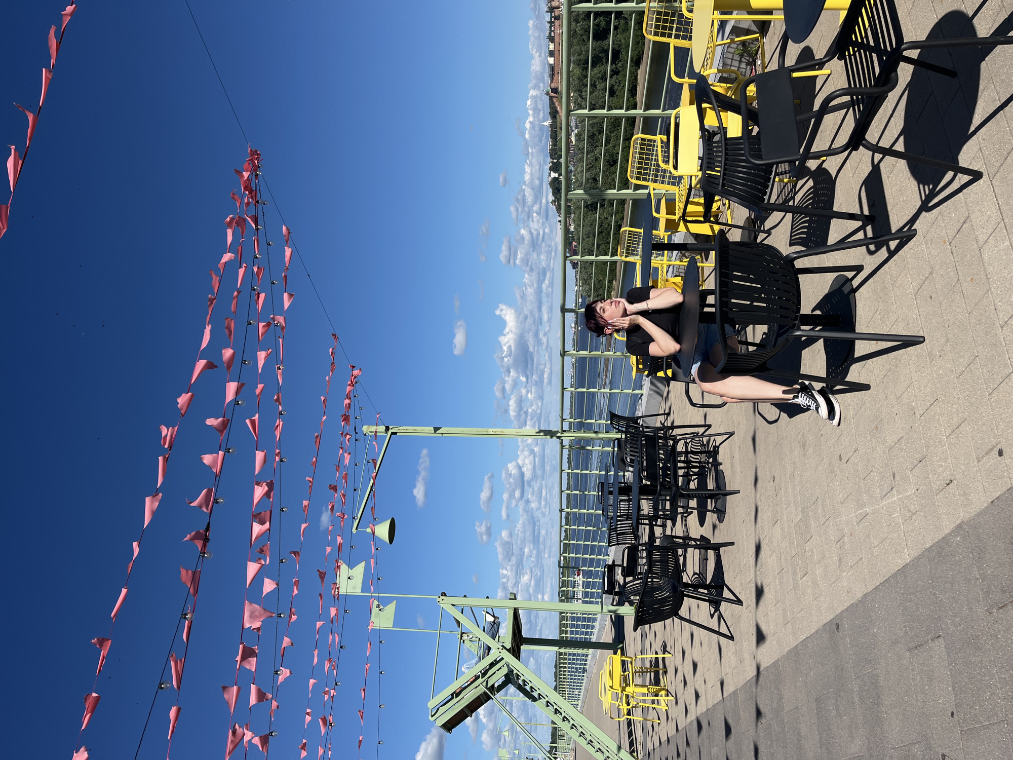 The image captures a sunny outdoor seating area with a view of a river and a cityscape. Pink pennant banners are strung across the top, with string lights interspersed. In the foreground, a woman with dark hair is seated at a black table, resting her chin on her hands and looking to the side. She is wearing a black t-shirt and denim shorts, with black and white sneakers. Around her are several black tables and chairs, with some bright yellow tables and chairs visible further back. A metal railing separates the seating area from the river. In the background, across the river, a city skyline with buildings and trees is visible, possibly indicating a location in Nizhny Novgorod, Russia, given the architecture and river setting. A green metal structure with stairs is also present on the left side of the image. The ground is paved with interlocking stones.