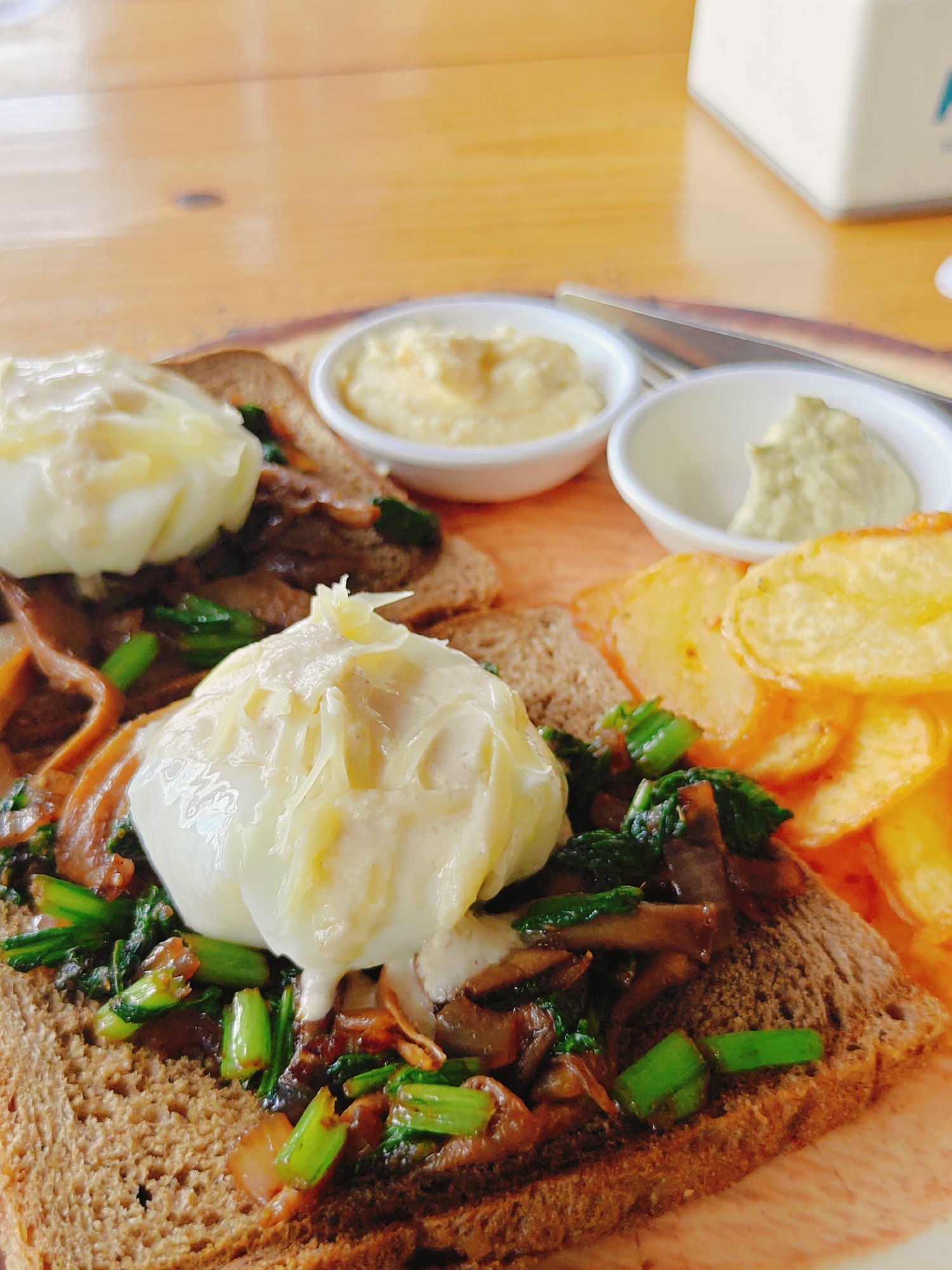 The image shows a close-up of a plate of food, likely a breakfast or brunch dish, presented on a wooden surface. The dish consists of two pieces of whole-wheat toast, each topped with poached eggs and a mixture of sautéed vegetables, possibly spinach and mushrooms, and what appears to be thinly sliced cooked meat. One of the poached eggs is partially obscured by the other. Beside the toast, there are two small white bowls containing dips or sauces. To the right of the bowls, a pile of golden-brown potato chips is visible. The background shows a blurred wooden table and a portion of a white object, possibly a napkin dispenser or a sugar dispenser. The lighting suggests an indoor setting, possibly a cafe or restaurant. The image does not provide any specific landmarks or clues to pinpoint its location within Pokhara, Nepal.