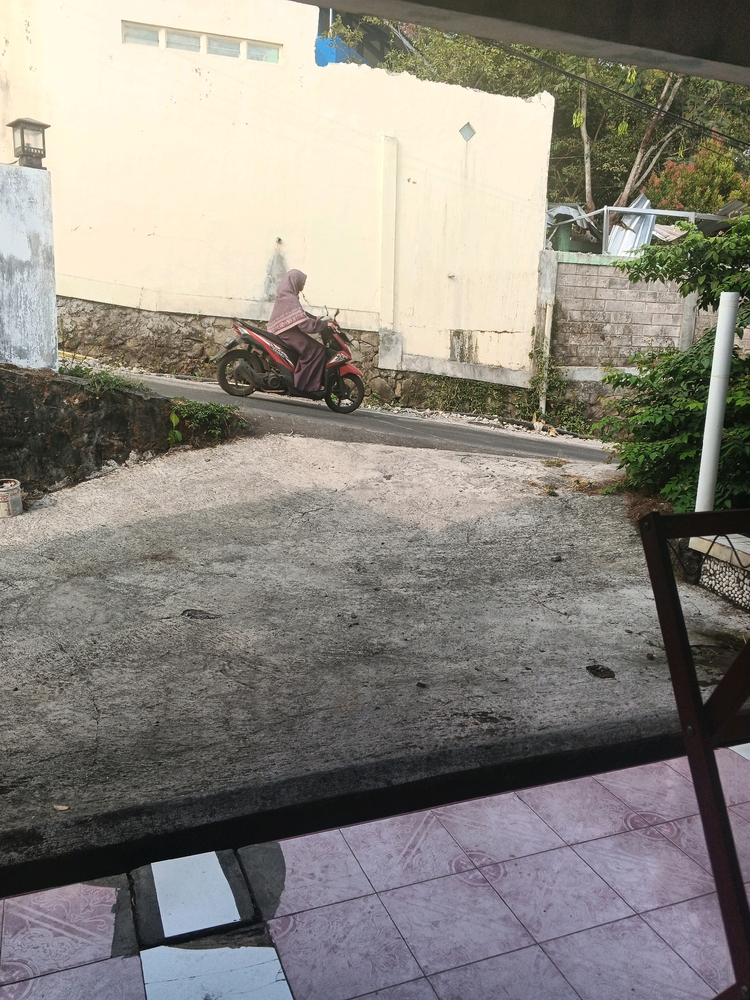 A person wearing a maroon hijab and a maroon dress rides a red motorcycle on a paved road. The motorcycle is moving from left to right. The person is facing forward and appears to be driving the motorcycle. The background consists of a cream-colored wall and a stone wall. There is vegetation and trees to the right of the scene. The foreground shows a concrete surface and a tiled patio. The scene appears to be an outdoor residential area, possibly in Keji, Indonesia.