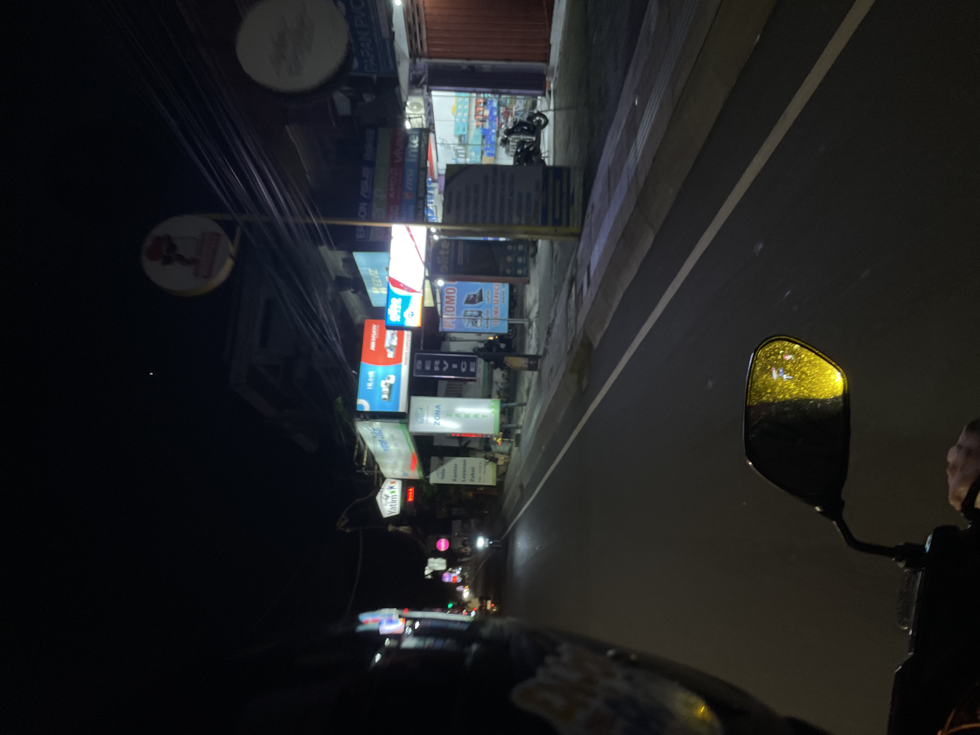 This image depicts a nighttime scene on a street in Denpasar, Indonesia, likely from the perspective of someone riding a motorcycle. The foreground is dominated by the dark silhouette of the motorcycle, with its wing mirror reflecting yellow streetlights and a hint of the rider's face.
The background shows a row of shops illuminated by various neon signs and light bulbs. Several signs are clearly visible with text, including "Yatim", "BURA", "Kantor Layanan Zakat", "ZONA", "Hlosk", "MAYIAW", "SERVIC", "EZVIZ", "ERON ASLES", and "intel". One sign also advertises "TERIMA SERVICE" and "PROMO". A motorcycle is parked outside one of the shops. The road surface is visible, with a white line marking the lane. Overhead, electrical wires crisscross the sky. The overall impression is that of a busy commercial street at night, with the artificial lights creating a vibrant but somewhat murky atmosphere. There are no strong indications of weather conditions beyond the reflections on the mirror which might suggest slight dampness. The scene is captured from a low angle, emphasizing the perspective of movement.