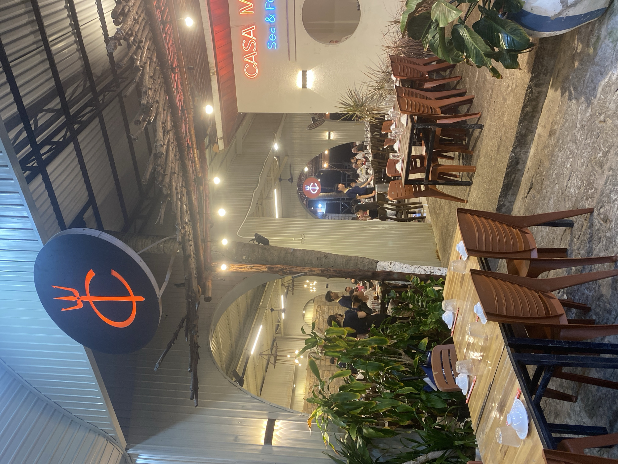 This is a photograph taken at night, showing an outdoor seating area of a restaurant in Mũi Né, Vietnam. The restaurant has a rustic, open-air design with a corrugated metal roof supported by metal beams and wooden elements. In the foreground, several wooden tables are set with white plates, glasses, and red chopsticks. Orange plastic chairs surround the tables. Lush green plants are visible to the left and right of the tables.
Further back, through an arched opening, more tables and chairs are set up, with people seated and dining. The restaurant's interior features hanging light fixtures and fans. A prominent circular sign with an orange trident logo and the letter "C" in orange neon is visible above the arched entrance. To the right, a white wall displays the restaurant's name in neon lights: "CASA M" in orange and "Sec & Fo" in blue. The overall ambiance suggests a casual dining establishment, likely a seafood restaurant given the "Sec & Fo" signage which likely refers to seafood. The presence of palm trees and the general outdoor setting are consistent with a coastal Vietnamese location like Mũi Né.