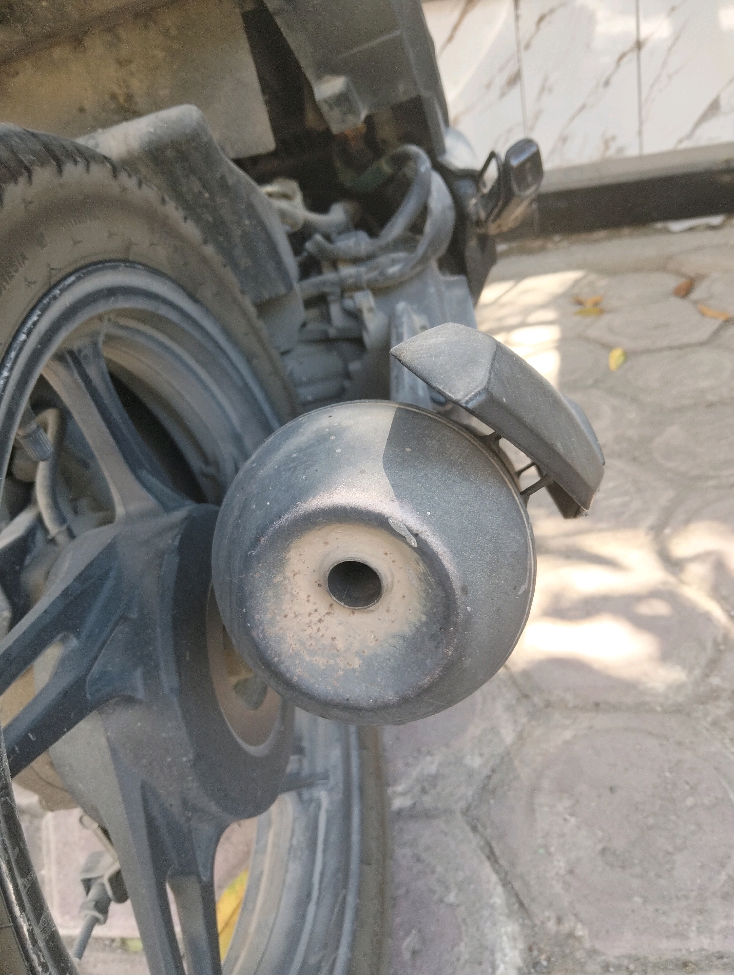 The image shows the rear wheel and exhaust pipe of a motorcycle parked on a paved surface, likely in Paciran, Indonesia. The motorcycle is dark gray and appears to be dusty, suggesting it has been used recently. The wheel has a black rim and a tire with visible treads. The exhaust pipe is round and metallic, also showing signs of dust and some reddish-brown discoloration, possibly rust or dirt accumulation. A dark gray plastic fender is visible above the exhaust pipe. The background consists of a textured wall with vertical lines and a paved ground with visible grout lines between the stones. Sunlight casts shadows on the ground, indicating an outdoor setting. There are no people or animals visible in the image, and no specific actions are being depicted other than the motorcycle being parked.