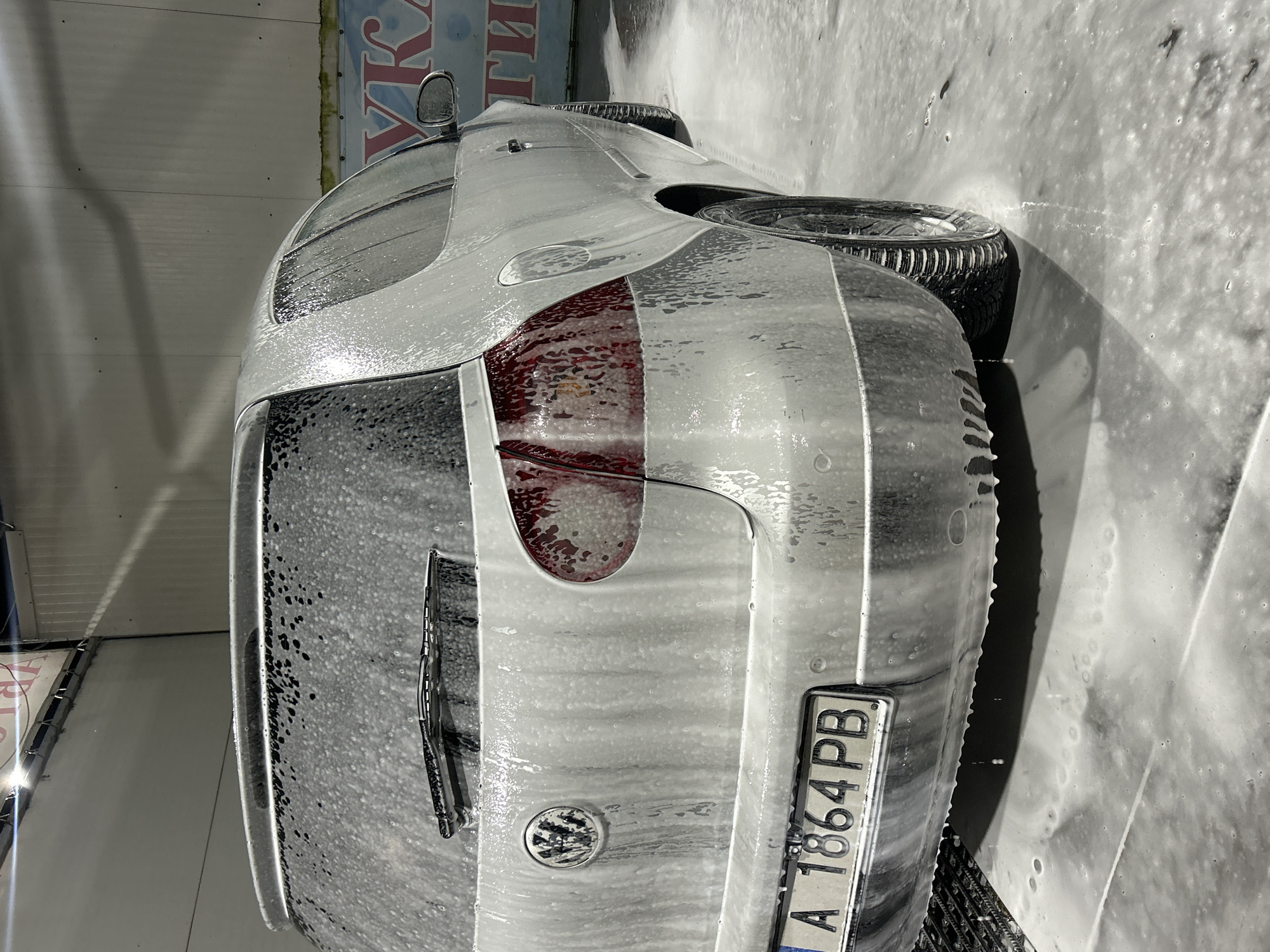 A silver Volkswagen Golf hatchback, likely a Mk5 model, is centered in a self-service car wash bay in Burgas, Bulgaria. The vehicle is thoroughly covered in white cleaning foam, which drips onto the wet, foam-covered concrete floor. The rear of the car is prominently featured, displaying a Bulgarian license plate "A 1864 PB," with "A" denoting Burgas. The Volkswagen emblem is visible on the tailgate, directly above the license plate. A rear windshield wiper is positioned at the top of the rear window, also coated in foam. The red taillights are visible on either side of the tailgate, with foam clinging to their surfaces. The rear bumper shows multiple circular parking sensors. The car's right side is partially visible, revealing the rear passenger door, side mirror, fuel cap, and a rear wheel fitted with a tire exhibiting distinct tread patterns. The interior of the car wash features light-colored paneled walls. Overhead artificial lighting reflects off the car's wet surfaces. A partially visible sign on the right wall displays blue and red Cyrillic letters, including "УКА" and "ТИ," consistent with a commercial establishment. No individuals are present in the image.