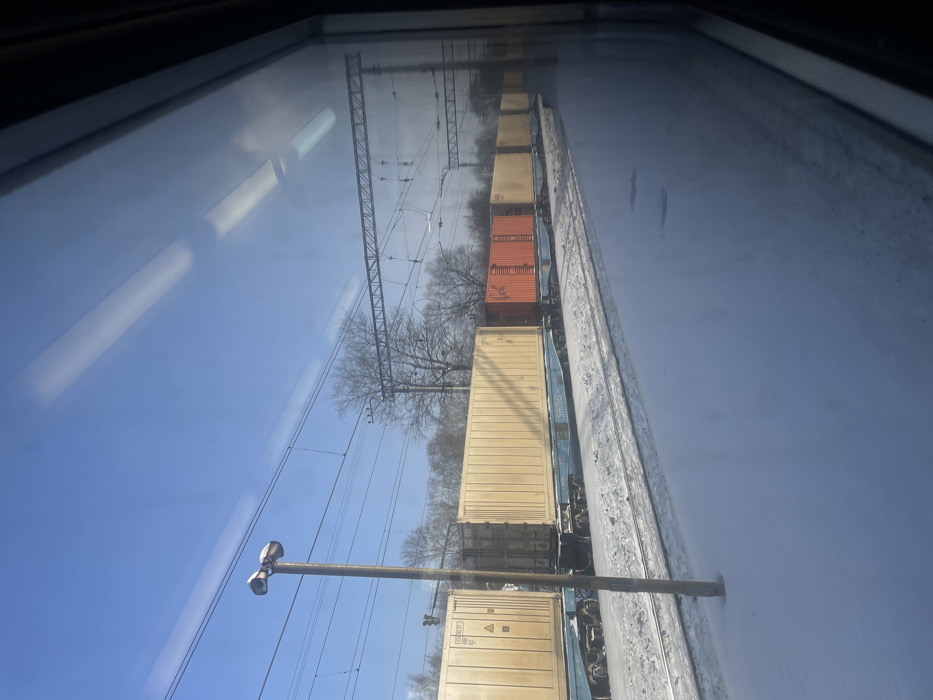 The image displays a snowy railway scene in Elektrougli, Russia, observed through a train window. A long freight train occupies a parallel track on the left, composed of multiple blue flatbed railcars. These railcars carry various intermodal shipping containers; the majority are light yellow or tan, with one prominent orange container situated in the middle of the visible train section. Markings "COH 957303" are discernable on a yellow container near the front, and the number "51097583" is visible on a blue railcar. The ground, including the tracks and an adjacent snow-covered platform or open area, is blanketed in white snow. A tall, light-colored lamp post with two top-mounted fixtures stands in the left-mid foreground. Numerous overhead electrical lines and metal gantry structures span the tracks against a clear blue sky. Deciduous trees, bare of leaves, line the distant background. Reflections of internal light fixtures are visible on the window pane. No individuals are present, and no actions are explicitly depicted.