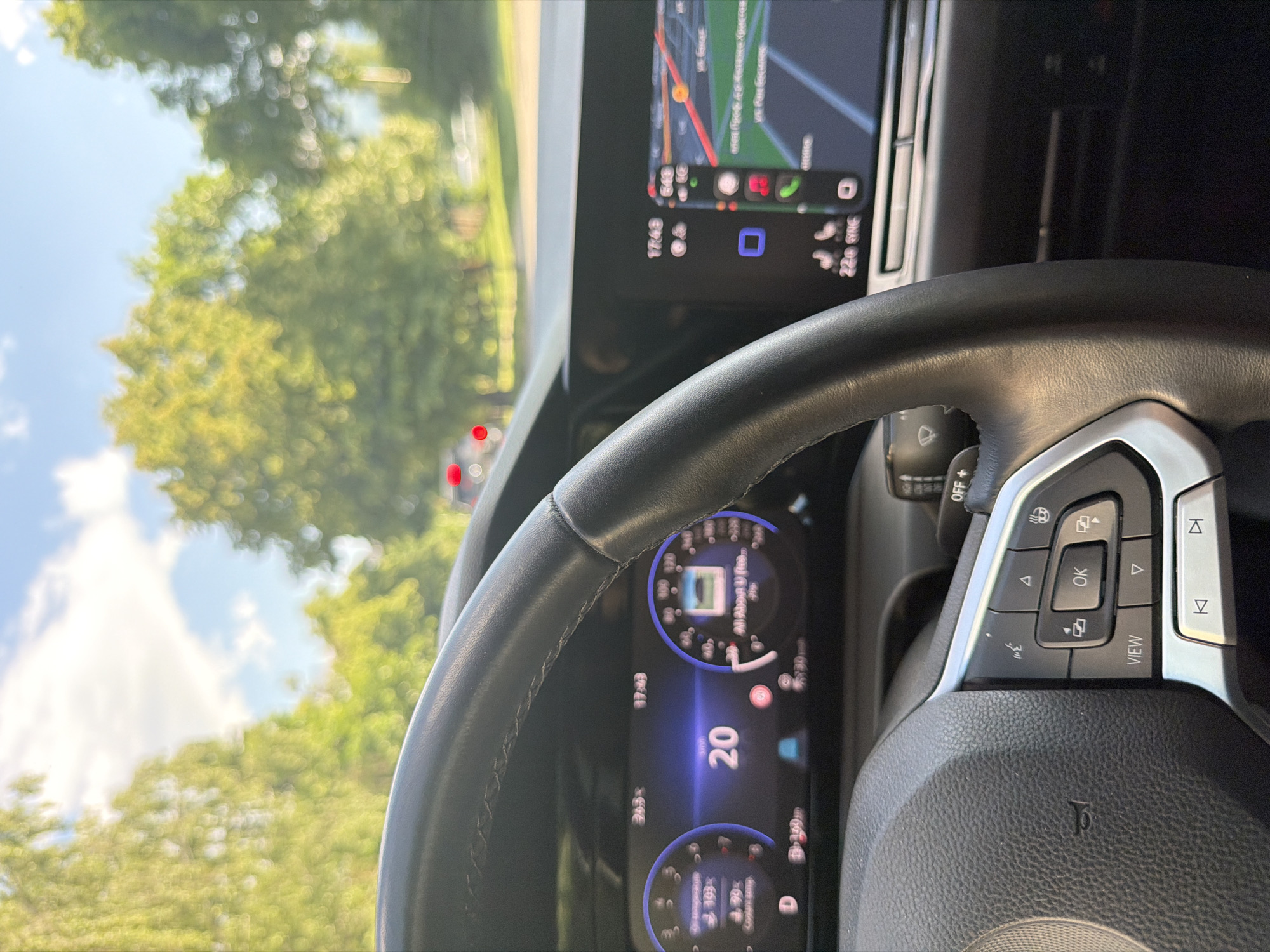 The image captures the interior of a car from a driver's perspective, rotated 90 degrees clockwise, looking out through the windshield and across the dashboard. The primary subjects are the car's steering wheel, digital instrument cluster, and infotainment screen, set against a blurred background of green trees and a bright blue sky with some white clouds.
The steering wheel, made of black leather with silver accents, is prominent in the foreground, partially obscuring the digital instrument cluster. It features various control buttons, including "OK," "VIEW," directional arrows, and icons for functions like heated steering wheel and wiper settings.
The digital instrument cluster, visible behind the steering wheel, displays a speed of "20" (likely km/h or mph). Other partially visible text includes "17:48" (the current time), "399.9" (possibly a trip meter), and what appears to be "Coolant Temp" and "All About U" on the circular gauges.
To the right, the infotainment screen shows a navigation map with various street paths and icons, suggesting active route guidance. This screen also displays "17:48" for the time and "22.0 SYNC," likely indicating the climate control temperature setting.
The outdoor scene, seen through the windshield, is brightly lit, indicating a sunny late afternoon, consistent with the "17:48" timestamp. The environment is lush with green trees. The car is located in Sofia, Bulgaria. The overall impression is one of being in transit or momentarily stopped in a vehicle.