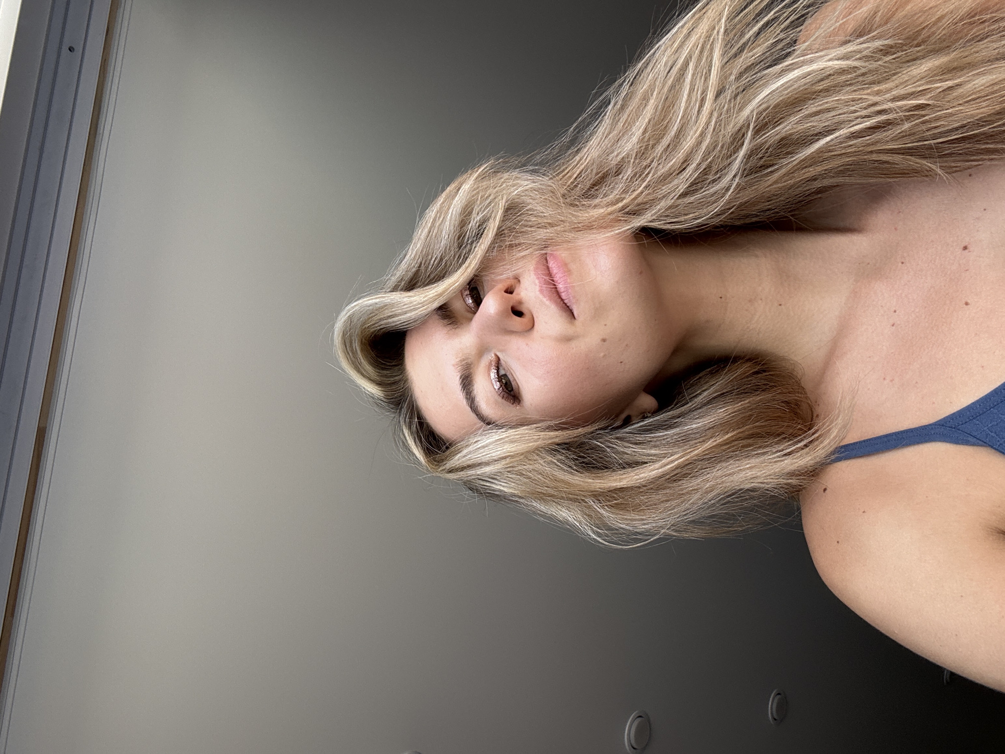 A woman with long blonde hair is facing the camera at an angle. Her hair is styled to one side, partially covering her face. She has brown eyes and fair skin with a few visible freckles. She is wearing a blue tank top. The background consists of a light gray wall with a window frame visible on the left side of the frame and a pair of ceiling lights in the lower half of the frame. The location is Unknown, Unknown.