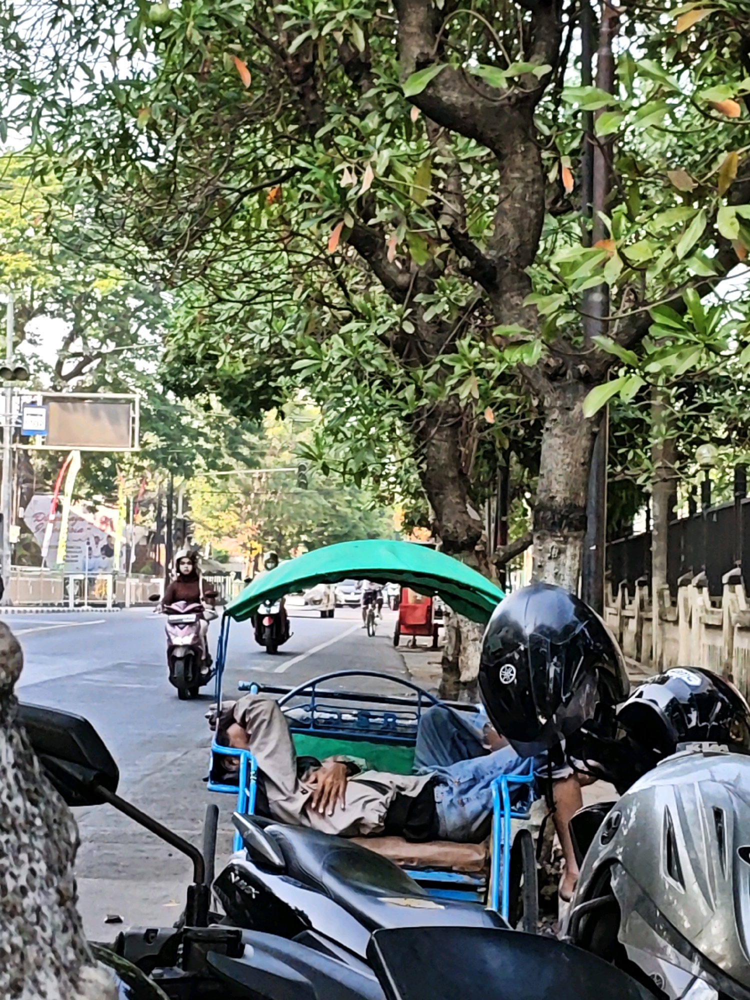The image depicts a street scene in Kediri City, Indonesia. In the foreground, several motorcycles are parked. One motorcycle's handlebar and a portion of its body are visible on the left. To the right, two helmets are placed on the seats of motorcycles. In the middle ground, a man is sleeping in the passenger compartment of a three-wheeled vehicle, which is covered by a green canopy. The vehicle has a blue frame and appears to be a becak, a type of pedicab. The man is lying on his back, wearing a light-colored shirt and blue jeans. Behind the becak, another becak with a red body is visible. On the street, a woman is riding a scooter, wearing a dark top and a white helmet. Another person on a scooter is seen in the background. The street is lined with trees and buildings. The overall scene suggests a typical day in an urban environment with various modes of transportation and pedestrian activity.