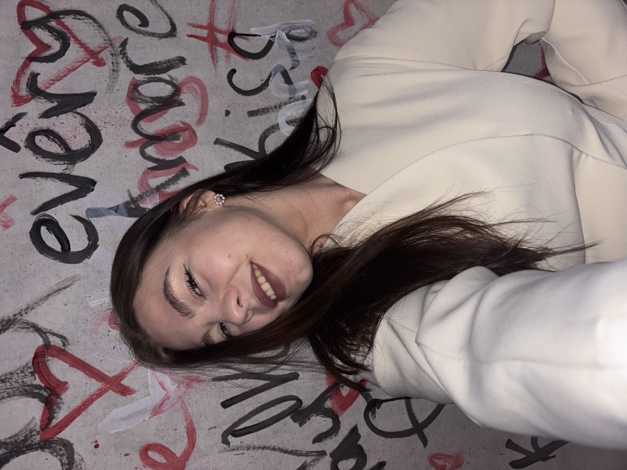 A woman lies on a surface with graffiti, smiling at the camera. The graffiti includes black and red paint in the form of writing, hearts, and lines. The woman has long brown hair, and she wears a light-colored jacket. She appears to be wearing a small, circular earring in her left ear. Based on the graffiti and overall style, the image likely originates from Perm, Russia.