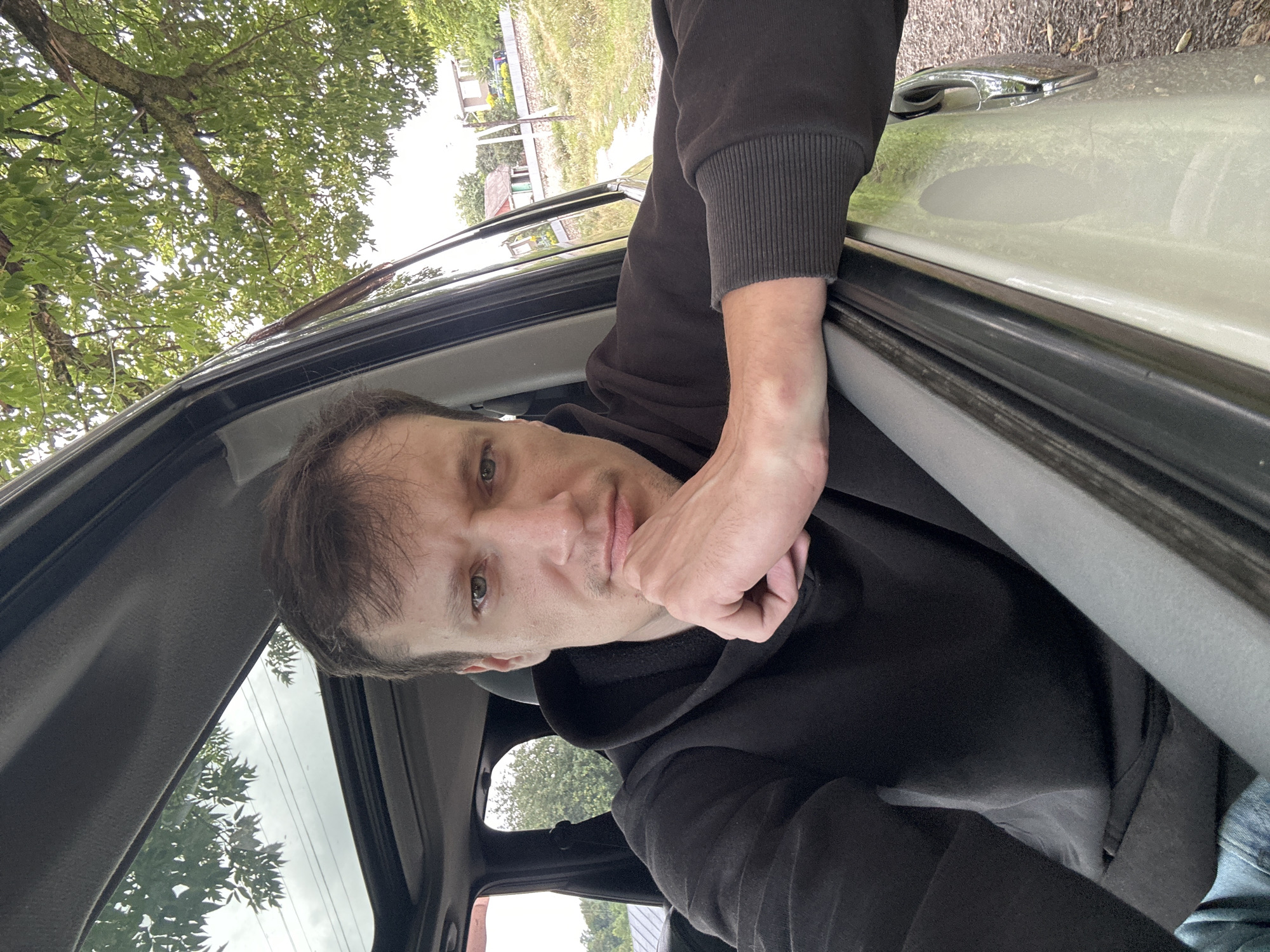 A man is seated in the driver's seat of a vehicle, looking towards the camera. His left arm is resting on the open car door, and his chin is propped on his left hand. He is wearing a dark hooded sweatshirt. The vehicle's sunroof is open, revealing green trees and a cloudy sky above. The background shows a rural setting with trees, a dirt road, and houses.