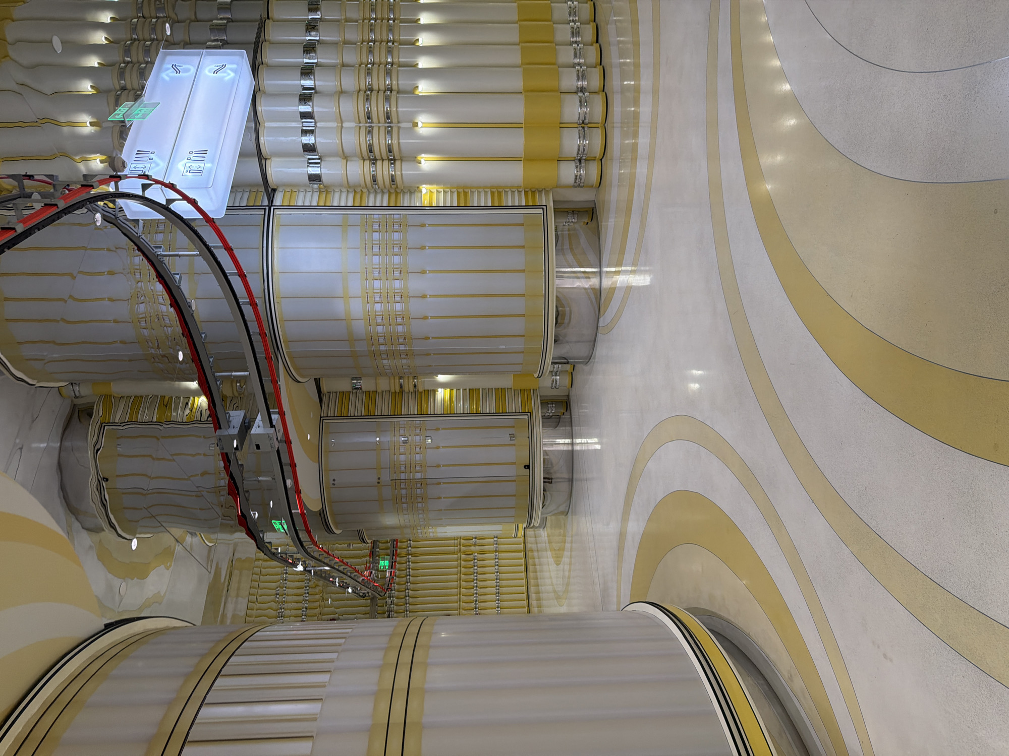 The image shows a top-down view of a modern interior, possibly a transportation hub or a large public building in Xuanwu District, China. The scene is dominated by large, cylindrical structures that run horizontally across the frame. These cylinders are white with yellow and silver detailing, creating a repeating pattern. Some of them appear to be part of a track or conveyor system, with a red and black cable track running above them.
Prominently featured is a brightly lit, rectangular object with glowing blue panels displaying white symbols. These symbols appear to be arrows indicating direction and perhaps information about different zones or functions. Two distinct areas, labeled with symbols that resemble simplified representations of people or figures, are visible on the illuminated panels. These panels are mounted on a curved track system.
The floor is decorated with large, flowing, curved patterns of yellow and white, suggesting a deliberate and artistic design. The lighting is bright and diffused, creating reflections on the smooth surfaces of the floor and the cylindrical objects. There are no people visible in the frame, but the presence of signage and the scale of the architecture suggest a public space designed for movement and interaction. The overall impression is one of advanced infrastructure and contemporary design, with a clean and futuristic aesthetic. The environment appears to be indoors and well-maintained, with no indication of specific weather conditions or time of day beyond the artificial lighting.