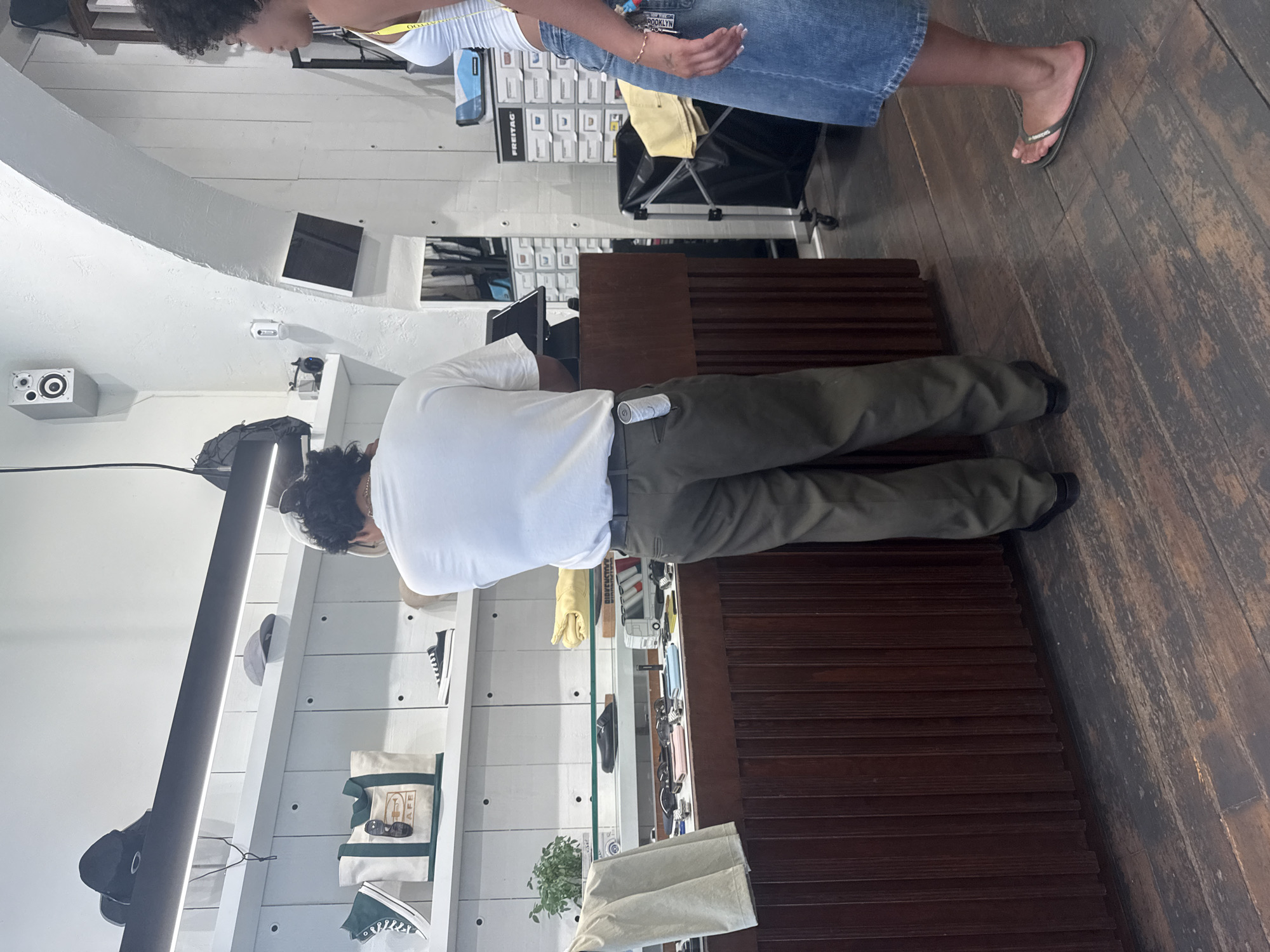 The image shows two people in what appears to be a retail store. On the left, a person with dark, curly hair is wearing a white tank top and denim shorts. They are standing and looking downwards. On the right, a person wearing a white t-shirt and olive green pants is bending over behind a counter, appearing to be attending to something on a shelf or display. Their back is mostly to the camera.
The store has white, slatted walls with various shelves displaying merchandise. Visible items include bags, sunglasses, and shoes. There is also a sound system speaker mounted on the wall. The floor is made of dark, distressed wood. A tall, dark wooden display unit is positioned to the right of the counter. The lighting suggests it is daytime. The overall impression is a casual, boutique-style retail environment.
A can of what appears to be a beverage is visible in the back pocket of the person in the green pants. Some text is visible on a bag on the shelf, reading "CAFÉ". Other text on merchandise or signage is difficult to decipher due to resolution or angle. The scene appears to be taking place indoors in a commercial setting.