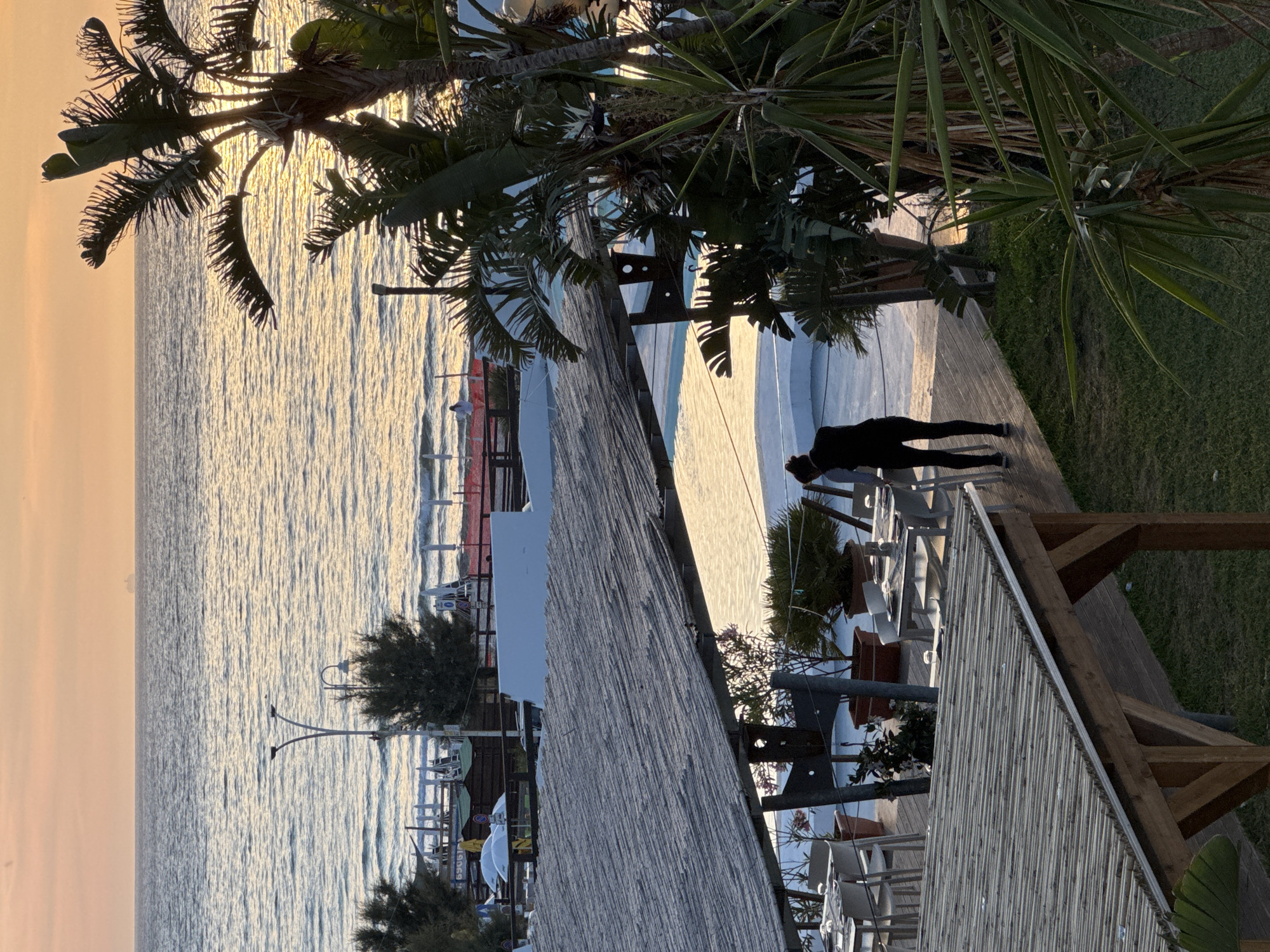 This image depicts a tranquil coastal scene in Isola delle Femmine, Italy, likely during sunrise or sunset, given the warm, golden light reflecting across the water. The sea occupies the left side of the frame, its surface rippled and shimmering with sunlight, extending to a soft, pastel-colored horizon. Along the distant shoreline, faint outlines of beach umbrellas and loungers can be seen.
In the foreground and mid-ground, a wooden deck runs parallel to the coastline, leading to a grassy area. A person, dressed in dark athletic attire, is visible on the wooden deck, appearing to be engaged in a stretching or exercise posture, possibly a plank or a forward bend. The area also features white outdoor tables and chairs, suggesting a dining or relaxation space.
Lush, dark green palm trees and other tropical foliage frame the top and right sides of the image, their leaves creating dramatic silhouettes against the bright, back-lit sky. Parts of a slatted wooden structure, possibly a roof or pergola, are also visible, adding to the architectural elements of the scene. A streetlamp stands near the water's edge, partially obscured by other structures. The overall mood is peaceful and serene, capturing a quiet moment by the sea. No discernible text is visible within the image.
