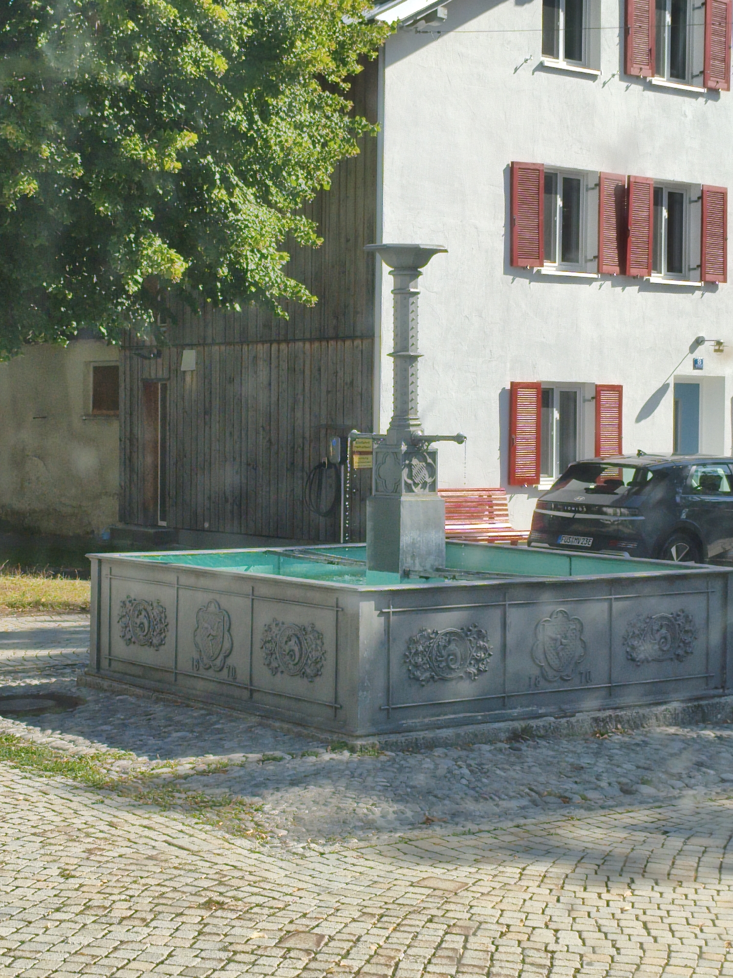 In Roßhaupten, Germany, a historic fountain stands in front of a modern building. The fountain is made of grey stone and has a large basin filled with turquoise water. Decorative crests adorn the sides of the fountain, along with the numbers "18" and "70." A tall, decorative pillar rises from the center of the fountain. To the right of the fountain, a dark grey car is parked. The building behind the fountain is white with red shutters on its windows. To the left of the building, a large tree with green leaves shades part of the scene. The ground in front of the fountain and building is paved with cobblestones.