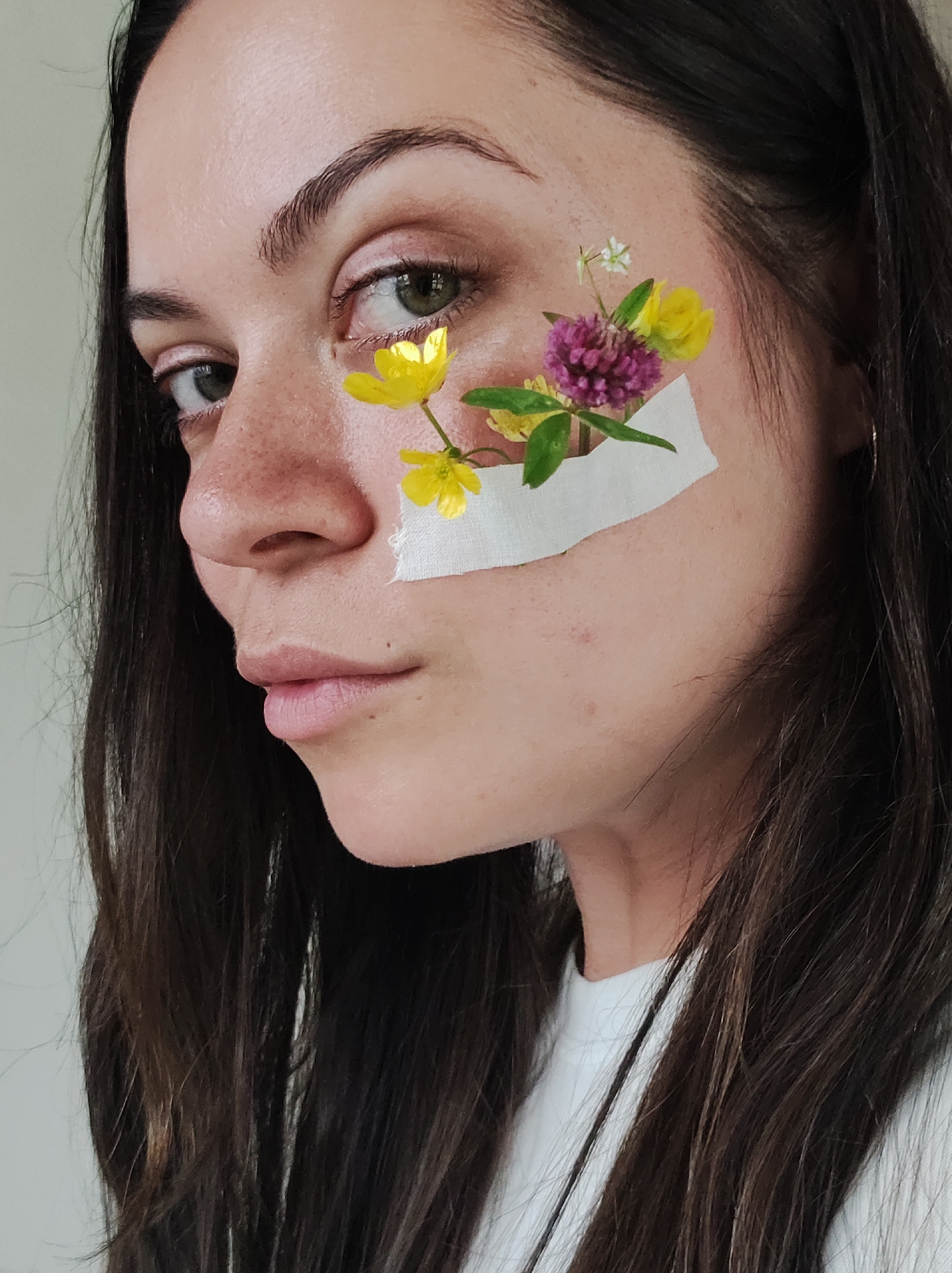 A close-up, profile view captures a young woman with long, dark hair, her face turned slightly to the left, revealing her left eye and cheek. Her skin is fair and dusted with freckles. A small arrangement of wildflowers, including yellow buttercups and a cluster of pink clover, is adhered to her cheek with a strip of white medical tape. The flowers are positioned near her eye, with some extending towards her cheekbone. Her makeup is subtle, with pinkish tones on her eyelids and lips. The background is a plain, light-colored wall. The lighting is soft and diffused, suggesting an indoor setting.