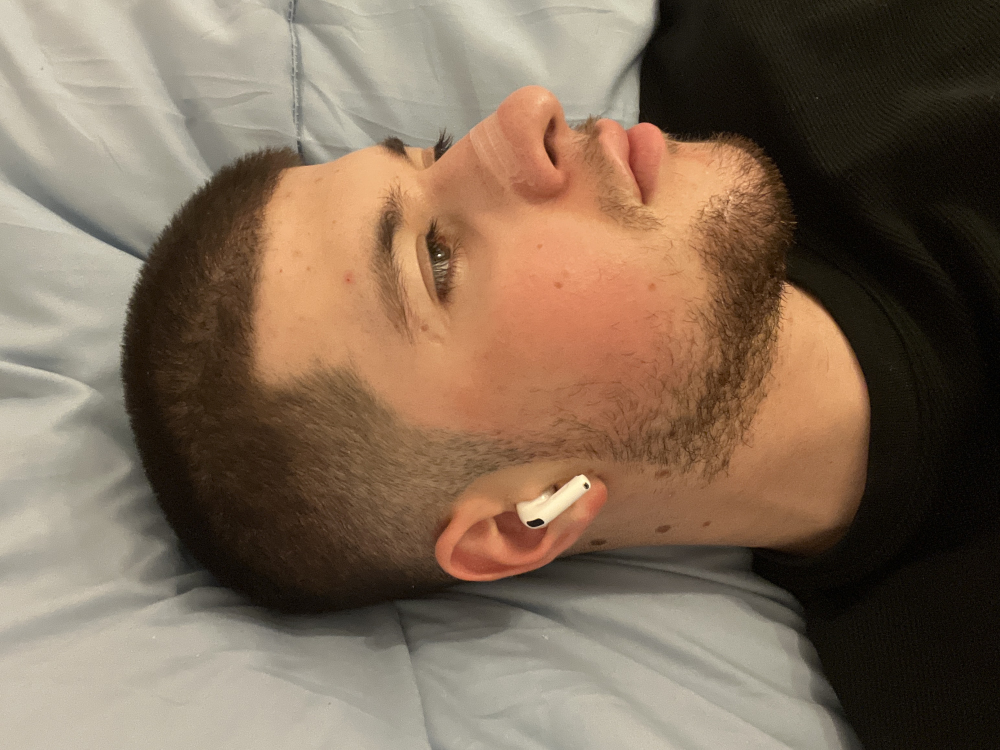 A close-up, side-view image shows a man lying on a light blue bed sheet, possibly indoors. He is light-skinned with short, dark hair and a beard. An earbud is visible in his left ear. The background includes a dark, possibly black, material, possibly clothing. The location is unknown, and cannot be deduced to be in Varna, Bulgaria, from the provided image.