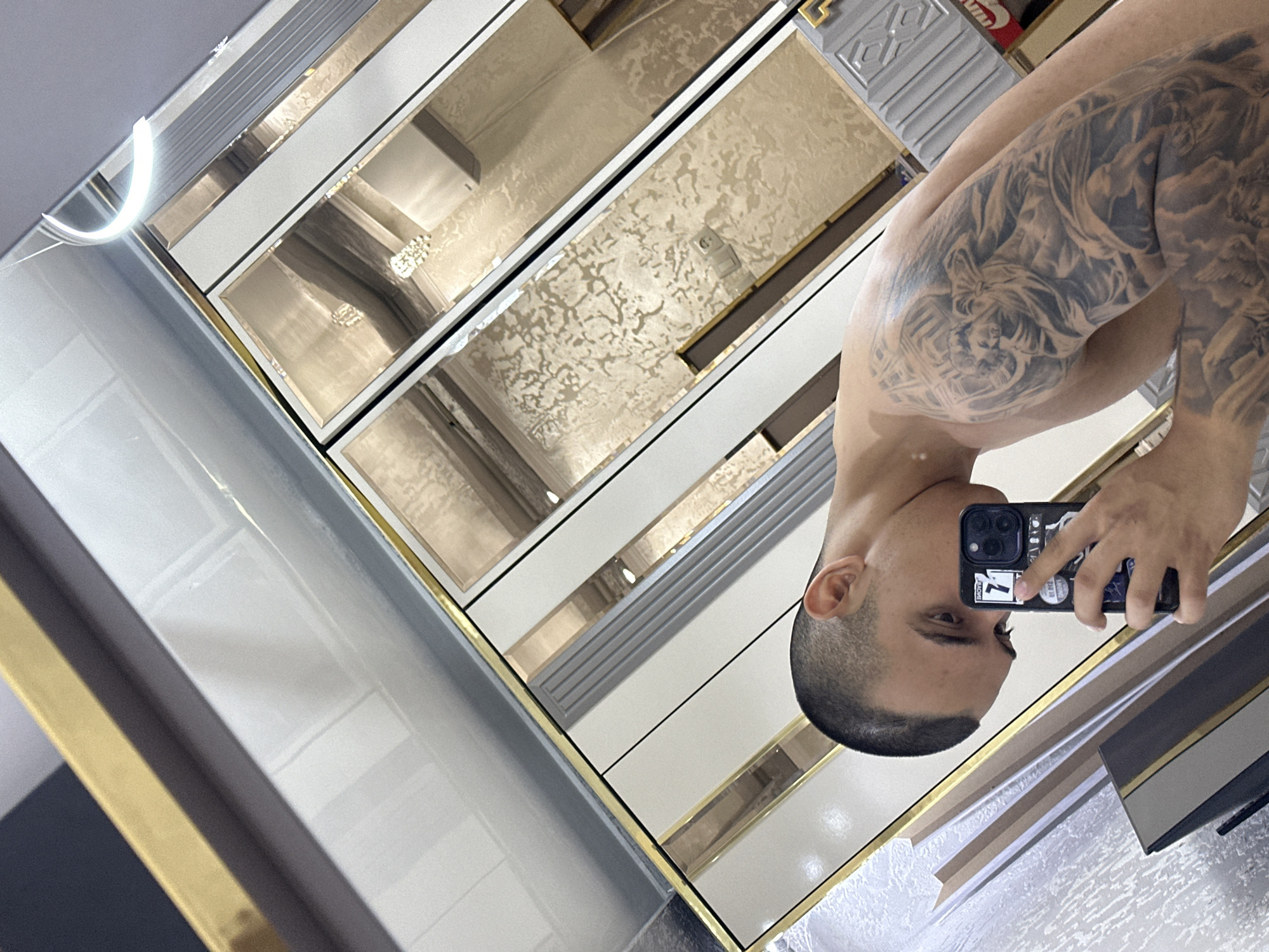 The image displays a mirror selfie of a man, captured from a rotated diagonal perspective. The man is reflected from the chest up, shirtless, showcasing a substantial, dark-toned tattoo on his right arm and shoulder. The tattoo artwork depicts detailed figures, possibly mythological or angelic, intertwined with clouds and intricate patterns. He has a short, dark buzz cut, and only the right side of his face, including his eye, is clearly visible, showing a neutral expression. He is holding a black smartphone with multiple camera lenses and various stickers on its case, including a clear "7" and a partial "LOVE" visible text.
The mirror is segmented, reflecting a modern interior design. It features alternating panels of a plain light color, possibly white or cream, and panels adorned with a textured, golden-beige pattern. These sections are bordered by thin, reflective gold-colored trim lines. Some areas also show light-colored panels with a ribbed or grooved texture. A bright, curved light source is prominently reflected in the upper left section of the mirror, suggesting indoor overhead lighting. The setting appears to be an upscale indoor environment, such as a stylish bedroom or living space. A small, indistinct red object with some white lettering, possibly a product label, is faintly reflected in the top right.