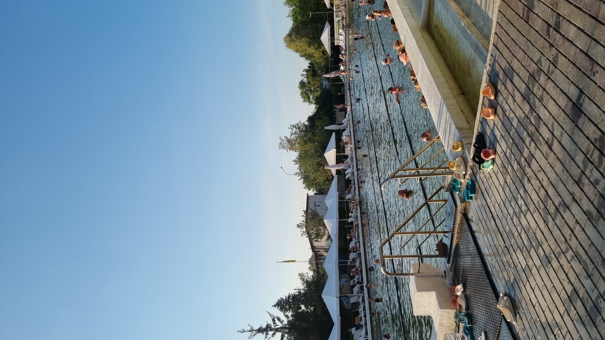 The image depicts an outdoor thermal pool facility in Berehove, Ukraine, under a clear blue sky. Numerous individuals are visible within the pool, some swimming and others standing. Along the poolside, lounge chairs are arranged under white canopies, with people relaxing on them. The foreground features a wooden deck with various flip-flops and sandals scattered around. The pool is bordered by concrete structures and vegetation. The overall scene suggests a warm, recreational day at a public swimming complex.
