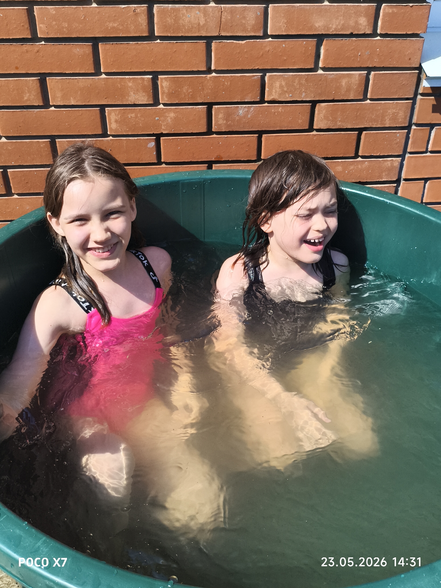Two young girls are sitting in a large, green plastic tub filled with water. The girl on the left is wearing a pink swimsuit with black straps that have white lettering, and she is smiling. The girl on the right is wearing a black swimsuit and has wet hair; she is also smiling and appears to be talking. The tub is placed against a red brick wall under bright sunlight. The lower left corner of the image shows the text "POCO X7" and the lower right corner shows "23.05.2026 14:31". The location is believed to be Chaikovsky, Russia.