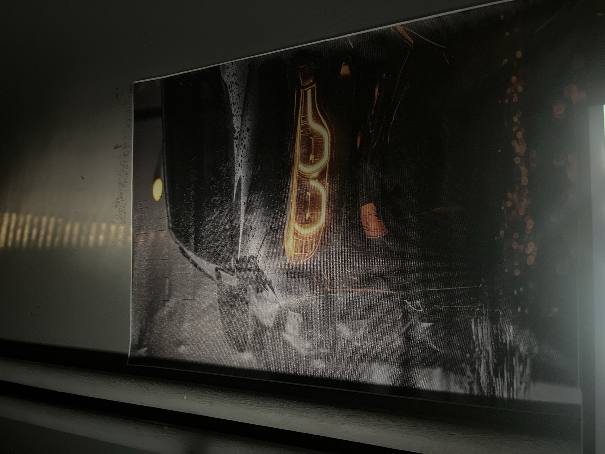 The image is a close-up shot of a framed canvas print hanging on a wall. The print depicts the front of a car, specifically the headlight, which is illuminated in an orange-yellow color. The rest of the car and background are rendered in grayscale, with visible textures suggestive of a canvas material. Water streaks are visible on the print. The location in Stara Zagora, Bulgaria cannot be determined from the image.