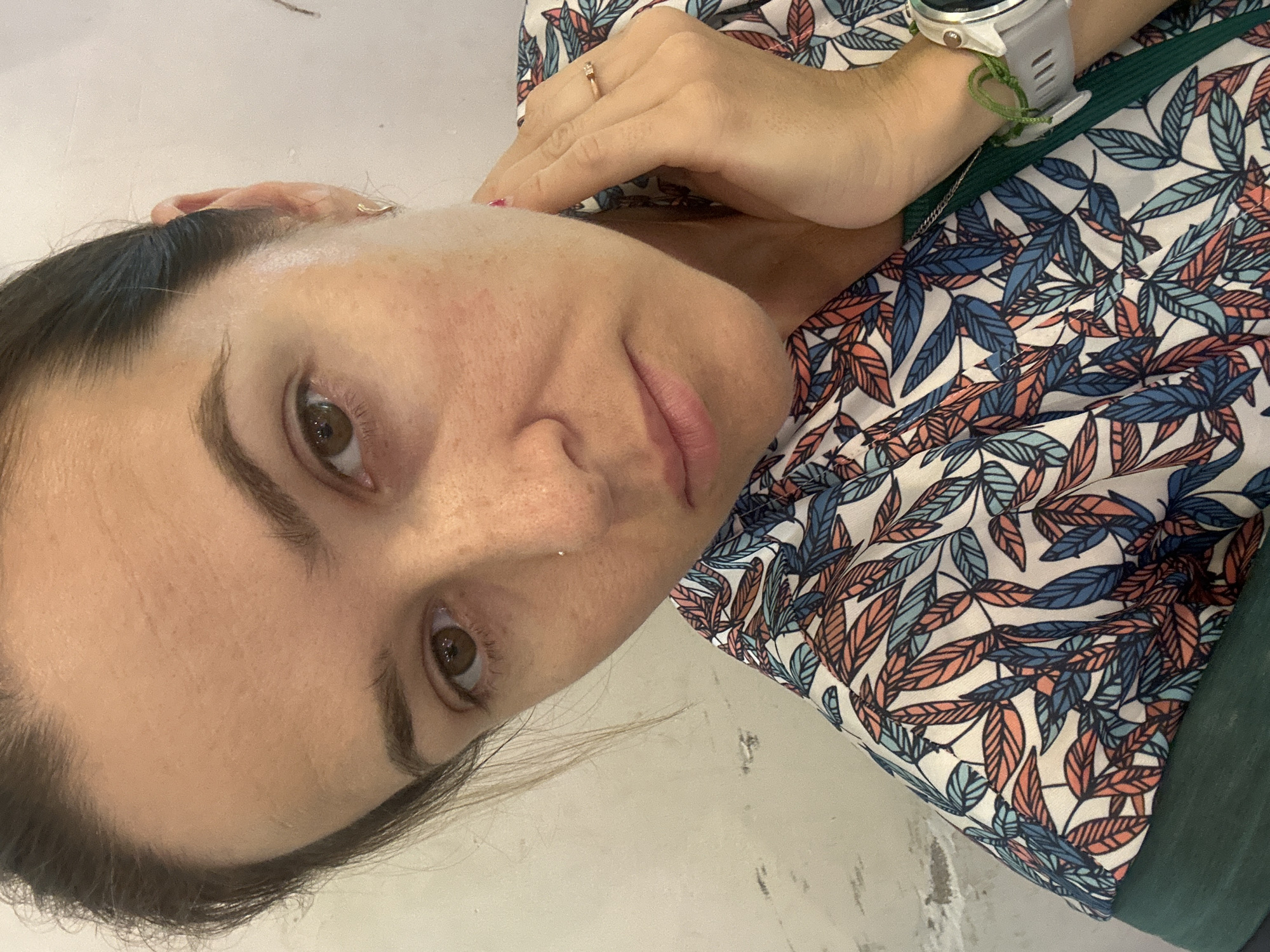 A medium shot captures a woman's face and upper body, angled slightly to the right, against a plain, off-white background. Her dark hair is pulled back, revealing her forehead and temples. She has brown eyes, arched eyebrows, and a small nose with a piercing. Her skin shows some texture and slight redness around the cheeks and forehead. Her lips are closed and slightly downturned, conveying a neutral or contemplative expression. Her right hand is raised to her neck, with a thin ring on her ring finger. She is wearing a patterned blouse with a design of colorful leaves in shades of blue, orange, and white. A dark green garment is visible beneath the blouse. A white smartwatch with a green band is worn on her left wrist. The scene appears to be indoors, given the lighting and the plain background. The location is not specified within the image itself.