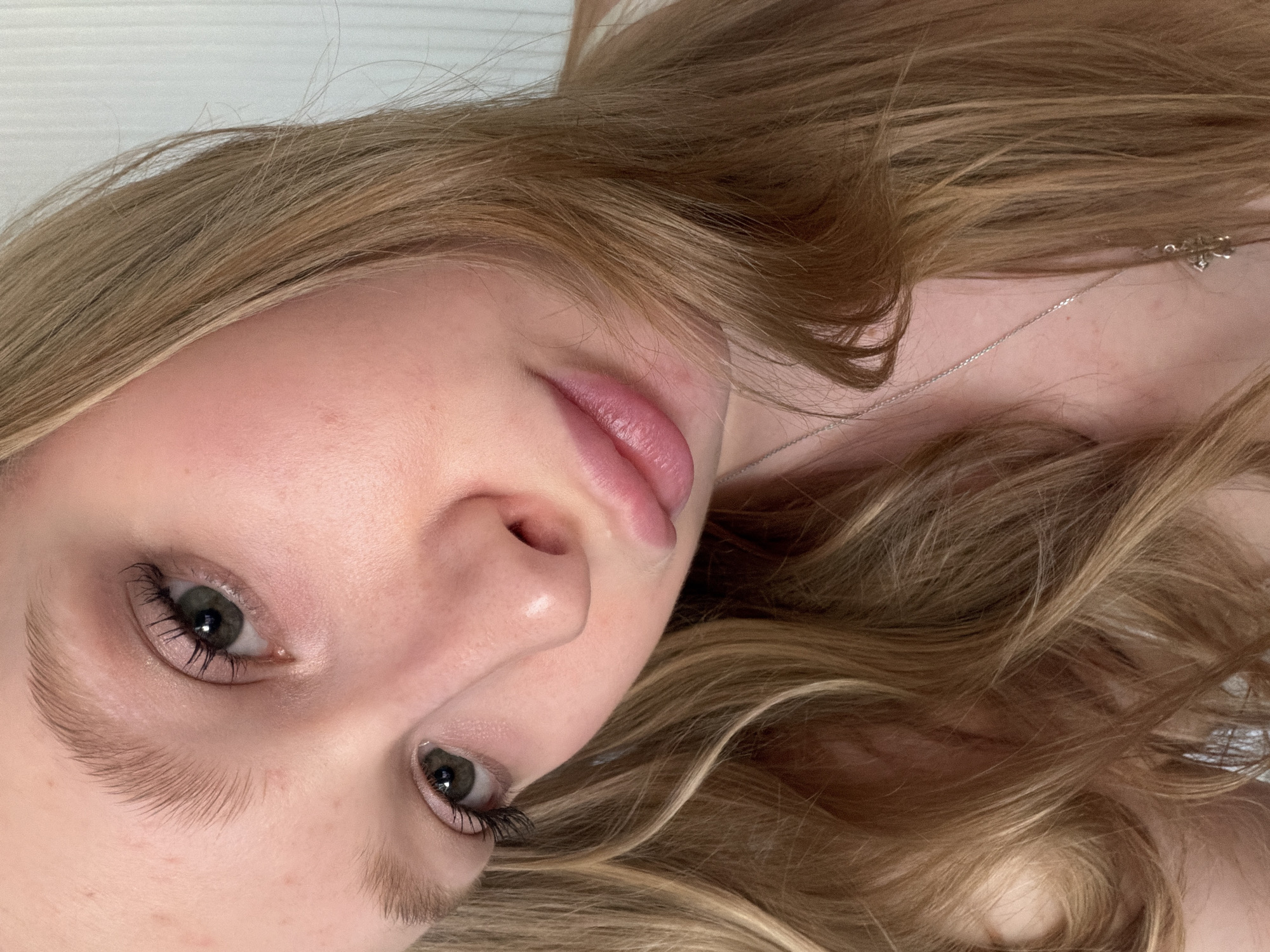 A close-up, angled portrait shows a young woman with light skin and light brown hair. She is facing upwards towards the camera. Her eyes are green and visible, and she has full lips. A silver necklace is visible on her neck. The background is plain and indistinct. The image was not taken in the city of Rossiyskiy, Russia.