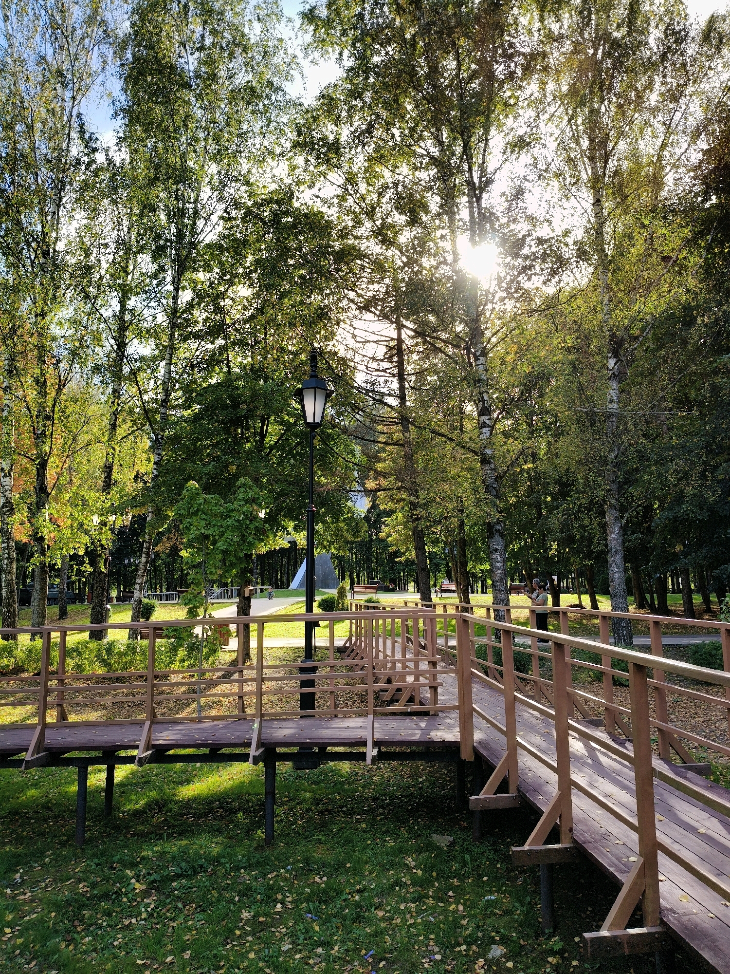 The image depicts a park scene in Naro-Fominsk, Russia, featuring a wooden walkway with railings winding through a wooded area. Sunlight filters through the green and yellowing leaves of tall trees, creating dappled light on the grass and walkway. A black lamppost stands beside the walkway, and a person is visible in the background walking on the path. Fallen leaves are scattered on the ground, suggesting it is autumn. The walkway is elevated in some sections, supported by dark metal posts. In the distance, beyond the trees, there are glimpses of park structures and benches.