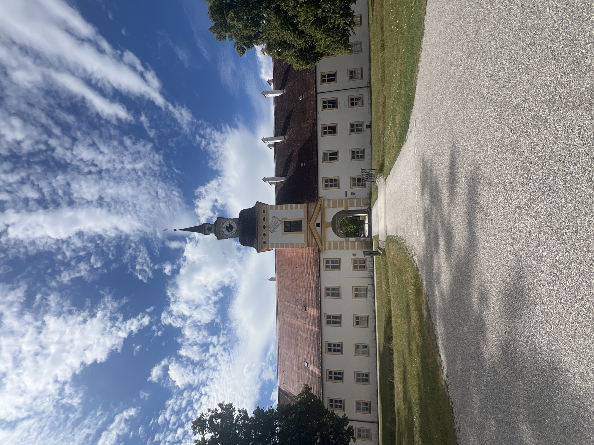 The image shows the south wing of Schleissheim Palace in Oberschleißheim, Germany. The building has a long, white facade with a red tiled roof and a central tower with a clock. The tower is topped with a spire. In front of the building is a gravel pathway leading to an archway under the tower. To the left and right of the pathway are grassy lawns. Trees are visible on either side of the building. The sky is blue with scattered white clouds. The image is taken from a low angle, looking up at the palace.