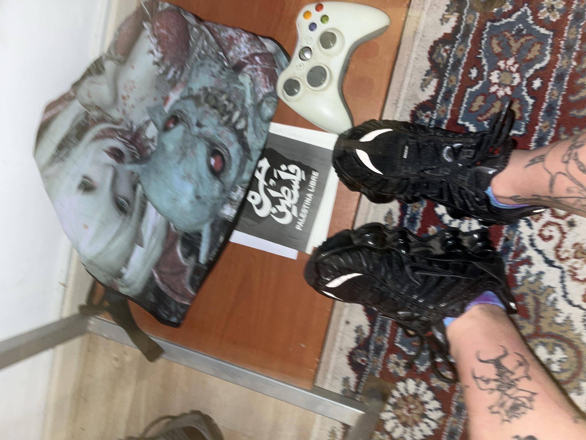 The image presents a top-down, slightly angled view of an indoor setting, featuring a person's lower legs and feet alongside several objects arranged on a patterned rug and a wooden surface.
**Subjects:** The primary subjects are the person's legs, visible from the mid-calf down, adorned with tattoos. They are wearing blue/purple socks and chunky black sneakers. Other objects include a backpack or bag with a striking graphic, a white Xbox 360 controller, and a book. The background comprises a multi-colored, ornate rug, a light brown wooden surface, and a white wall or baseboard.
**Activity:** The scene suggests a moment of rest or casual interaction within a personal space. The person's feet are placed on the rug, and the items are laid out around them.
**Notable details:** Both legs display distinctive tattoos. The right leg (from the viewer's perspective) features a design of a horned, possibly demonic, creature intertwined with a rose. The left leg has more abstract or script-like tattoos. The backpack exhibits a morbid artistic design, depicting two faces: one pale, human-like with red stains, and another blue-green, red-eyed, reminiscent of a zombie or monster. The white Xbox 360 controller shows some wear.
**Visible text:** On the book, prominent text in Arabic script reads "فلسطين حرة," which translates to "Free Palestine." Below this, the Spanish equivalent, "PALESTINA LIBRE," is also clearly visible. A partial "NIKE" logo is visible on the sole of the right sneaker.
