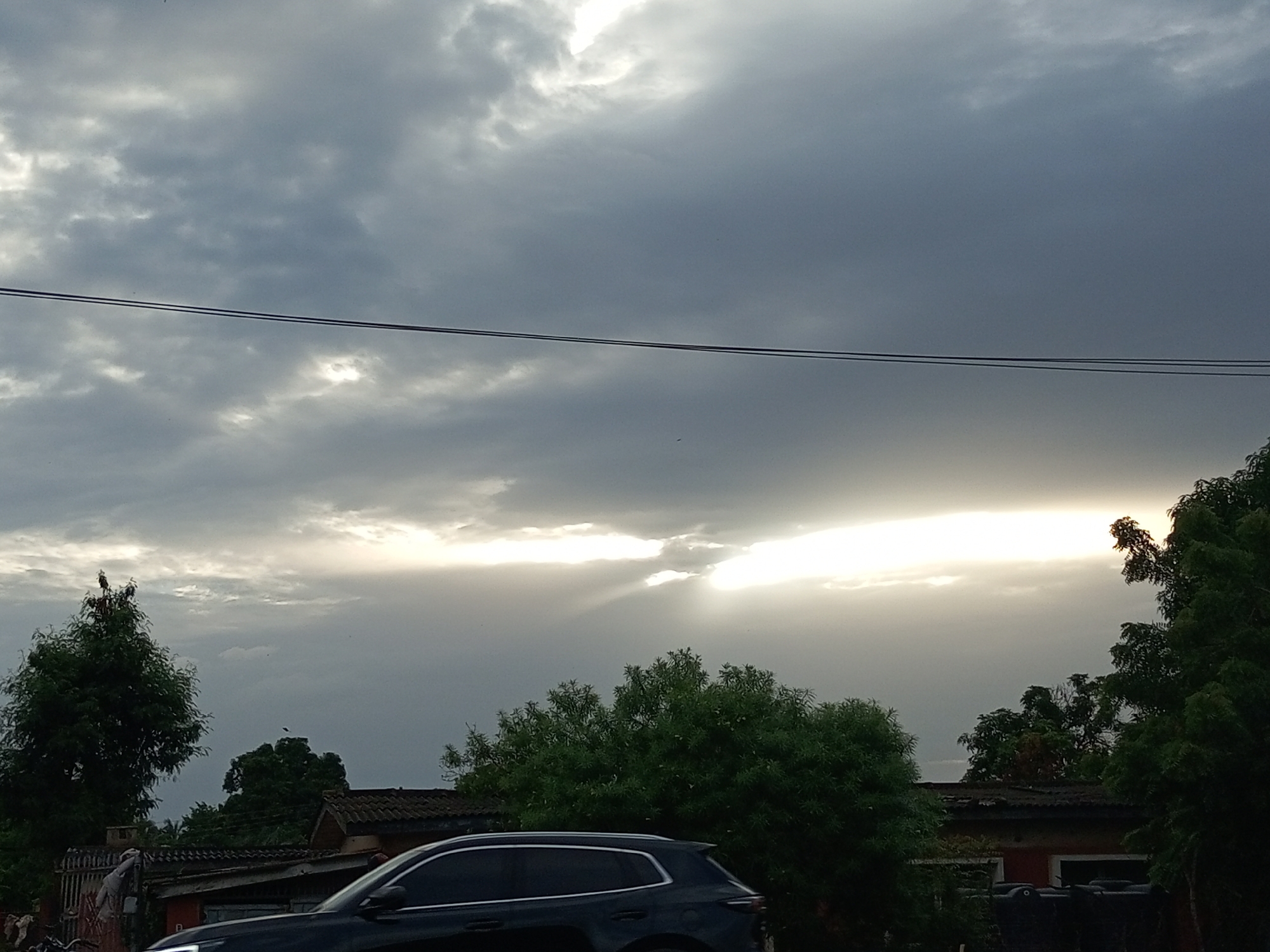 In Barden Yana, Nigeria, a dramatic sky dominates the scene with heavy, dark clouds partially obscured by a bright ray of sunlight breaking through, casting ethereal beams downwards. Below the tumultuous sky, two parallel power lines stretch horizontally across the frame. A lone bird is perched atop a tall, leafy tree on the left side of the image. The foreground features a dark-colored SUV, its tinted windows reflecting the muted light. Behind the vehicle, a collection of buildings with tiled roofs are visible, interspersed with lush green trees. The overall impression is one of impending weather or the aftermath of a storm, with a sense of natural power juxtaposed against the human infrastructure.