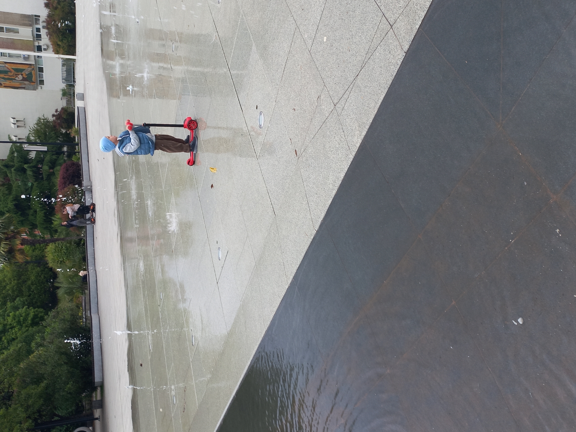 A child wearing a blue hat, vest, and brown pants is riding a red scooter in a public area. The area appears to be a square or plaza with a water feature, possibly a fountain, as water sprays upwards from the ground. The surface is paved with light grey tiles, some of which are wet. Adjacent to the tiled area is a darker, possibly wet, surface, potentially part of the water feature itself. In the background, there are trees and a building with multiple windows and some architectural details, perhaps a civic building or apartment complex. There are people walking and standing near the fountain. The photo was taken in Sochi, Russia.