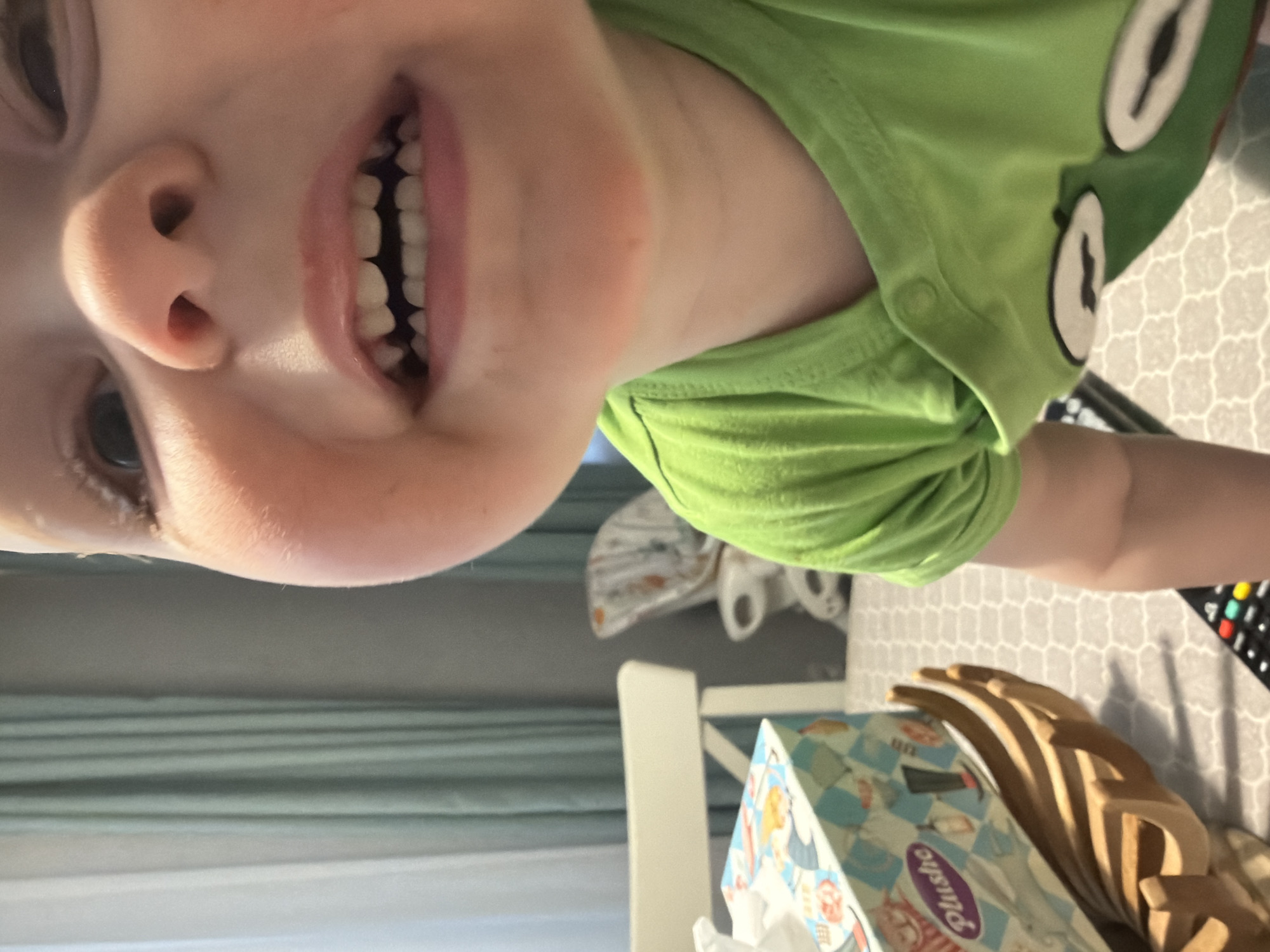 The image captures a close-up, upward-angled view of a young child, likely a toddler, smiling broadly. The child's face, predominantly the nose and mouth, fills the central frame, revealing a full set of upper and lower primary teeth. The eyes are open and looking slightly upward. The child wears a bright green short-sleeved t-shirt with a visible snap button closure on the right shoulder and a partial graphic, possibly depicting cartoon eyes, on the lower chest.
The background shows an indoor residential setting in Votkinsk, Russia. To the left of the child, a light blue and white patterned tissue box labeled "Plushe" rests on a light-colored surface, presumably a table. Next to the tissue box is a distinctive wooden object shaped like a hand. Partially visible in the lower foreground is a black remote control with colored buttons. In the deeper background, a white dining chair is present, and further back, a white high chair with a patterned seat cushion can be seen. Light teal curtains hang against a light-colored wall. The floor features a light-colored geometric pattern. The scene is brightly illuminated, with direct light emphasizing the child's face.
