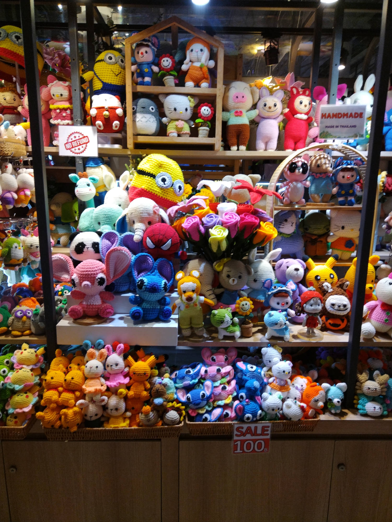 This is a full-frame, eye-level, medium shot of a store display filled with numerous handmade crocheted stuffed toys in Patong, Thailand. The toys are arranged on multiple shelves and in baskets, creating a colorful and whimsical display. The background consists of a wooden shelving unit with some hanging decorative elements. The store appears to be a souvenir or craft shop.
The subjects are primarily crocheted stuffed animals and characters. Prominently featured are numerous cartoon characters like Stitch, Elmo, Minions, and Hello Kitty, as well as generic animal designs such as rabbits, bears, elephants, and pigs. Many of the toys are brightly colored, with reds, yellows, blues, and pinks dominating the scene. Some of the toys are small and appear to be keychains or small trinkets, while others are larger plush toys.
There are no people visible in the image. The action is static, consisting of the arrangement of the merchandise. The environment is an indoor retail space, likely a market or a small shop, characterized by the wooden shelves and the dense display of toys. Two signs are visible: one reads "NO REFUND" with a circular red stamp, and another indicates "SALE 100" with a price possibly in Thai Baht. A "HANDMADE MADE IN THAILAND" sign is also visible, indicating the origin of the products. The lighting is somewhat diffused, typical of indoor retail environments. The overall impression is one of abundance and variety, showcasing a wide range of handmade crocheted items.
