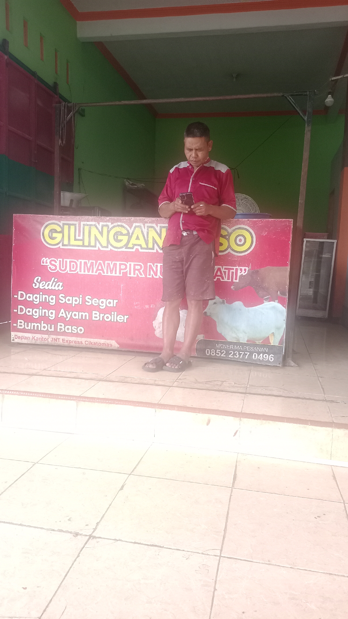 A man stands outside a shop in Karyamandala, Indonesia, looking at his phone. He is wearing a red and white polo shirt and brown shorts. Behind him, a red banner advertises "GILINGAN SO" and lists items for sale: "Sudiampir Nu," "Sedia," "Daging Sapi Segar," "Daging Ayam Broiler," and "Bumbu Baso." The banner also shows images of cows and states "MENERIMA PESANAN 0852 2377 0496." The shop appears to be a butcher shop. The ground in front of the shop is tiled.