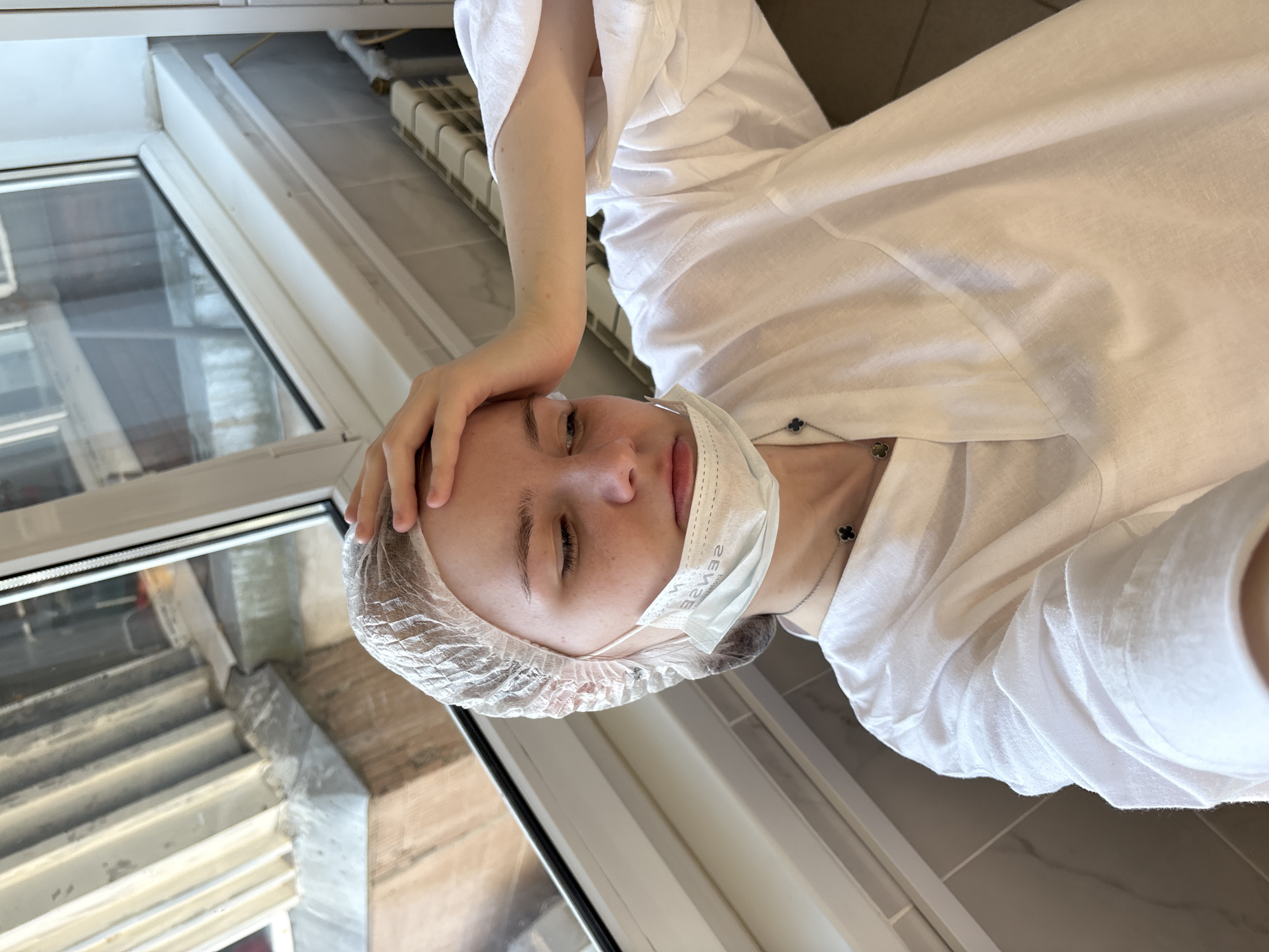 The image, rotated 90 degrees counter-clockwise, depicts a young person indoors, likely in a medical, cosmetic, or spa setting. The main subject is a light-skinned individual, possibly female, seen from a high angle. They are wearing a disposable white hair net, a white t-shirt or smock, and a light blue disposable face mask that is pulled down to cover their mouth but not their nose. Their eyes are closed, and one hand rests gently on their forehead. A silver necklace with a small black clover-shaped charm is visible around their neck.
The setting appears to be a room with a large window. Through the window, an urban exterior is visible, showing concrete structures and what look like steps or parts of another building. Inside, a white window frame, a white window sill, and a light-colored tiled floor are present. A radiator or heating unit is partially visible near the top right of the frame. Natural light streams in through the window, indicating it is daytime.
The person's closed eyes and the hand on their forehead could suggest they are resting, relaxing, or undergoing a treatment. The attire of a hair net, mask, and smock reinforces the idea of a clinical or beauty-related procedure. The text "SENSE" is clearly visible on the disposable face mask, appearing inverted due to how it's printed. The overall mood conveyed is calm, possibly reflective or restful. The location is an unknown indoor environment within Russia.