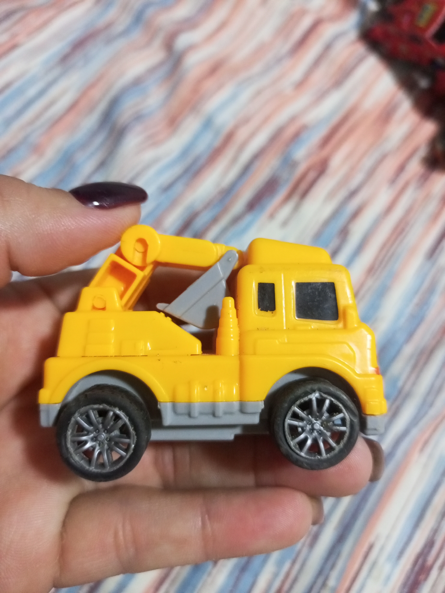 A person's hand with dark nail polish holds a small yellow toy tow truck. The truck has a grey crane arm and grey wheels with silver rims. The background is a blurry pink, blue, and white striped fabric. The image is taken from a slightly overhead angle, focusing on the toy truck and the hand. The location of La Tagua, Venezuela is not depicted in the image.