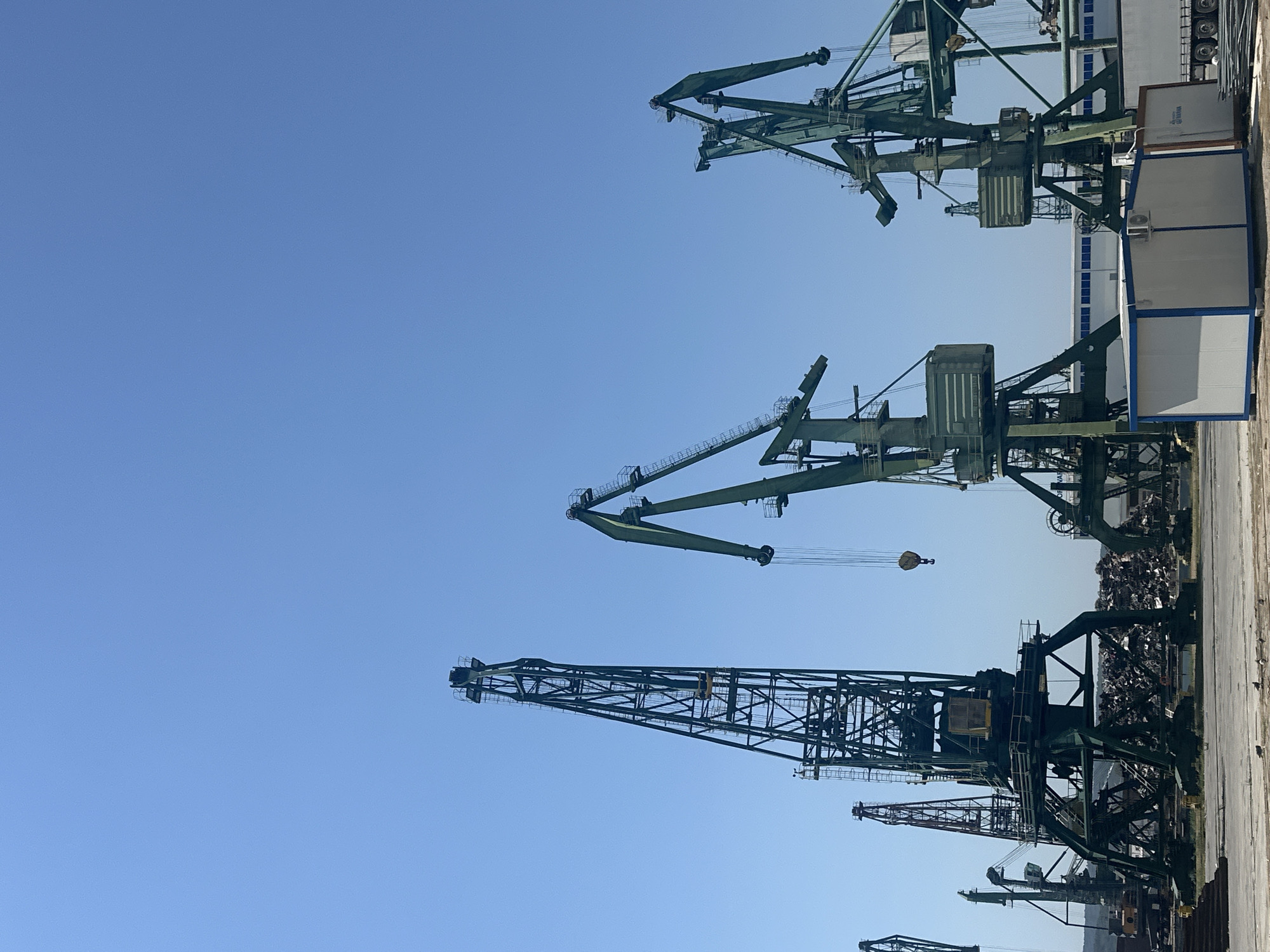 The image depicts a sunny day at a port, with a clear blue sky dominating the upper half. Several large, dark green industrial cranes are the primary focus, their towering structures silhouetted against the sky. One crane on the far left is a tall, lattice-style structure. To its right, a massive, more modern-looking crane features a prominent boom and counterweight. In the center, another large crane with a similar design is visible. To the right, two more cranes, also dark green, are positioned. In the foreground, a pile of scrap metal or debris is heaped behind the base of a crane. A small, white portable building with blue trim is situated in the lower right quadrant, with a portion of a white truck and some metal sheeting visible nearby. The ground is paved or concrete. The overall impression is one of industrial activity at a harbor or scrapyard.