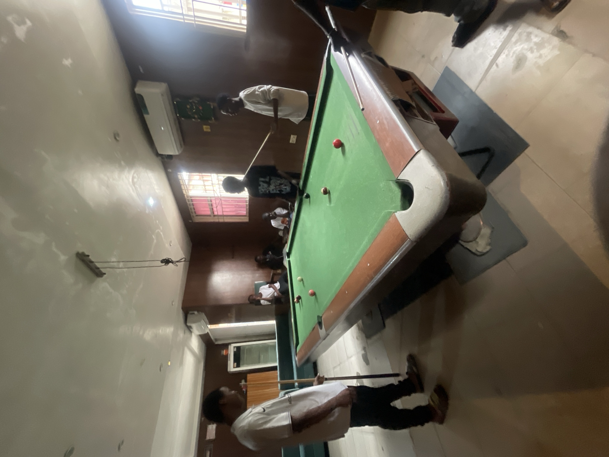The image depicts an indoor scene, likely a recreation room, with a pool table as the central focus. Four young Black males are present, engaged in a game of pool. One individual, wearing a white t-shirt and black pants, stands to the left of the pool table, holding a cue stick. Another, in a black t-shirt with white lettering, is positioned behind the table, also with a cue stick. A third person, dressed in a white long-sleeved shirt and dark pants, stands opposite the first individual, holding a cue. The fourth person, in a white t-shirt and black pants, is seated in a chair to the right of the table. The room has brown wooden walls, a green pool table with several colored balls, and large windows with red-tinted blinds. An air conditioning unit is mounted on the wall. The flooring appears to be tiled. The lighting suggests it is daytime. This scene is situated in Akowonjo, Nigeria.