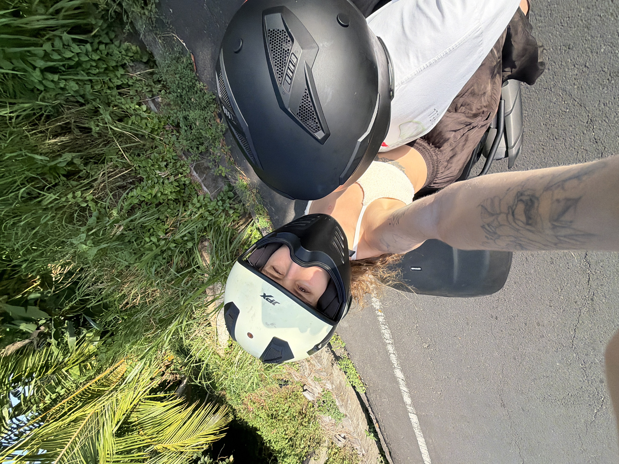 This image shows a person wearing a white and black helmet with "JPX" branding, looking at the camera. They are wearing a light-colored tank top and brown pants. Another black helmet is visible to their right. The person's tattooed arm is in the foreground, reaching out. They appear to be sitting on a scooter, with a paved road and lush green vegetation in the background. The scene suggests a location in North Kuta, Indonesia, likely on a road with abundant tropical foliage. The sunlight indicates it is daytime.